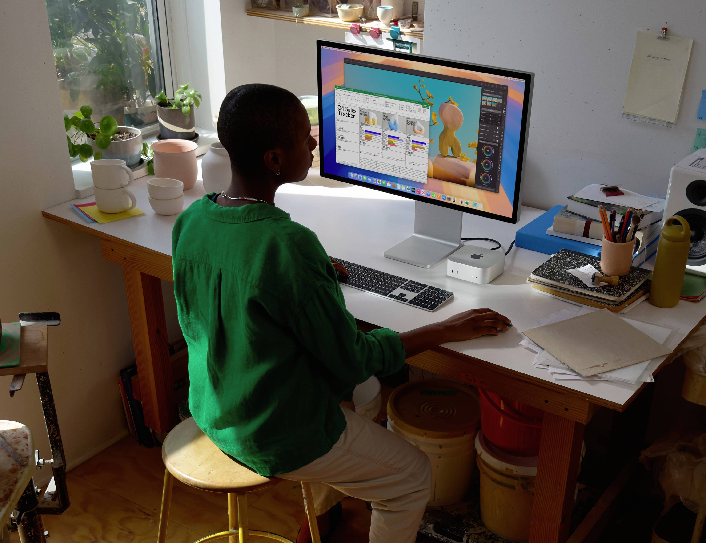 Eine Person sitzt an einem Schreibtisch und arbeitet an einem Computer mit einem Designprogramm auf dem Bildschirm, umgeben von Künstlerbedarf und Pflanzen.