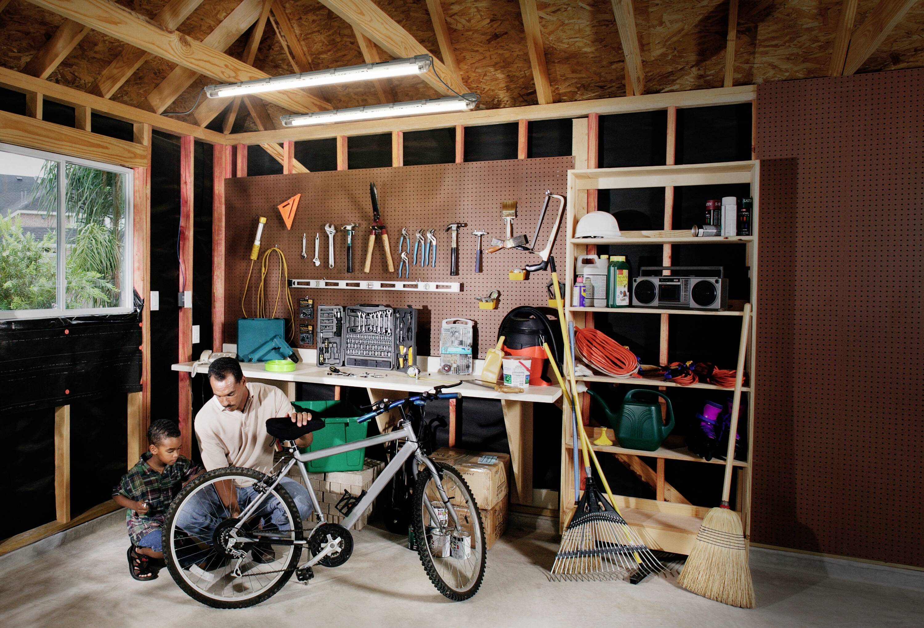 In einer Werkstatt repariert ein Mann mit einem Kind ein Fahrrad. Hinter ihnen hängen Werkzeuge an der Wand.