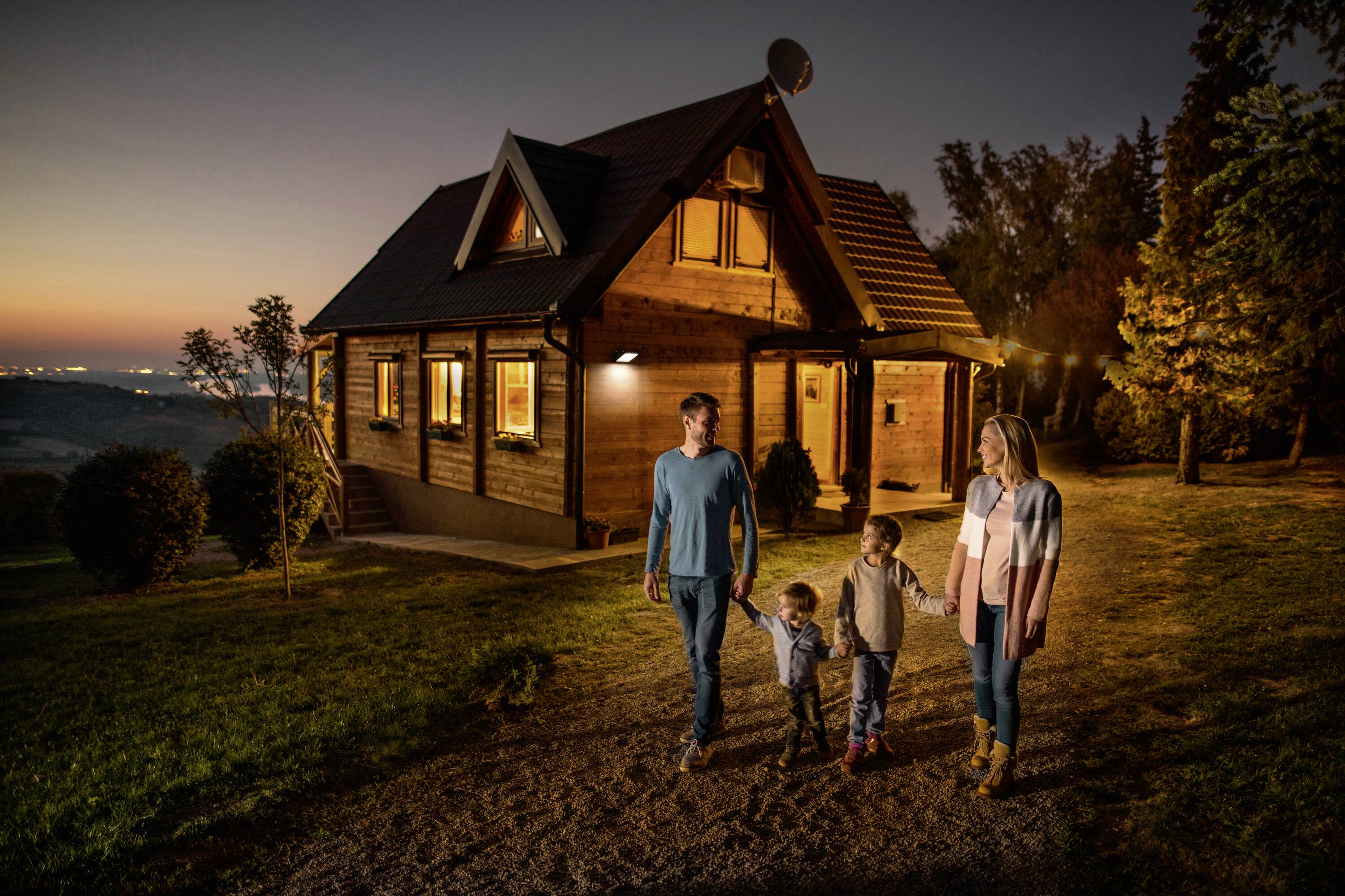 Eine Familie spaziert am Abend vor einem beleuchteten Holzhaus in ländlicher Umgebung. Der Himmel zeigt ein sanftes Abendrot.