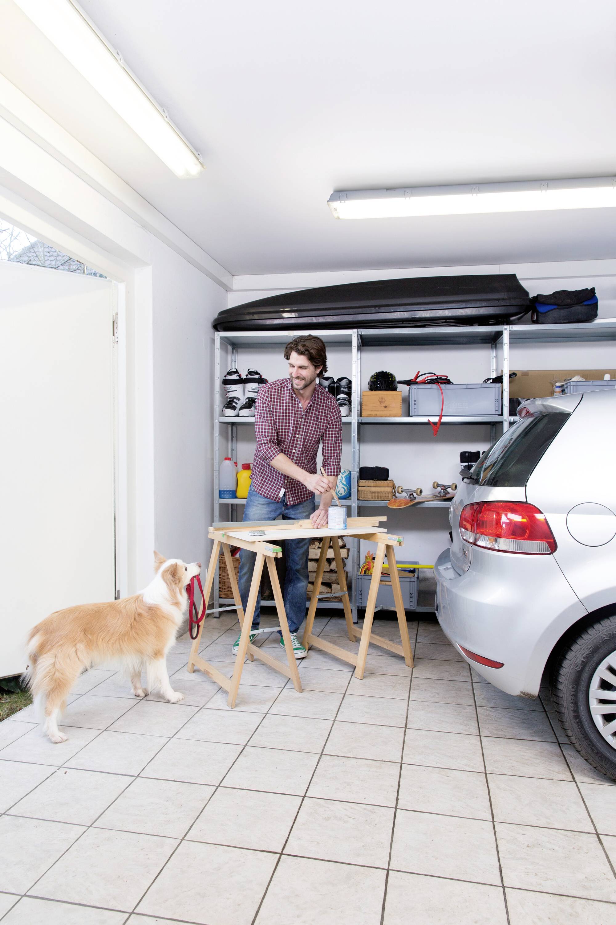 Ein Mann in einer Garage arbeitet an einem Werktisch. Ein Hund steht neben ihm. Im Hintergrund sind Regale mit Werkzeugen und Autoteilen.