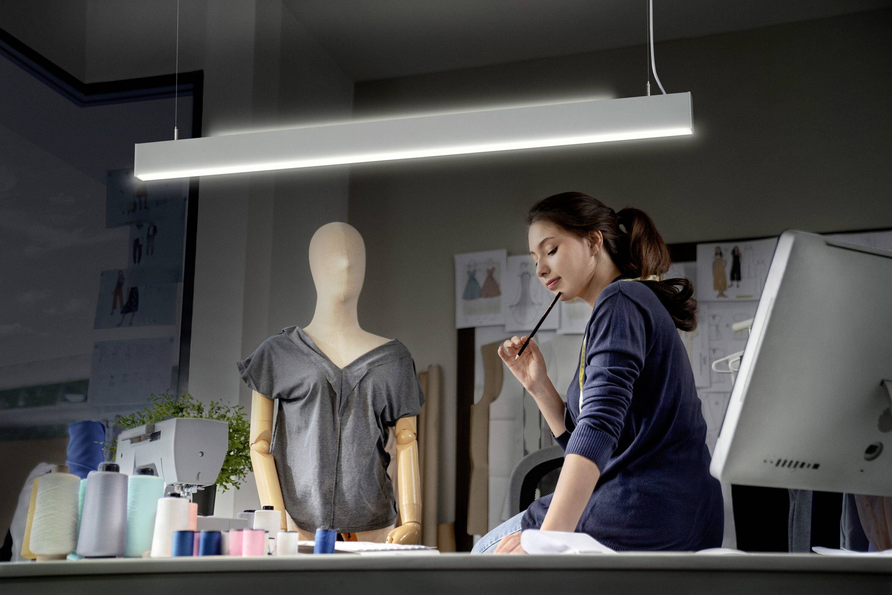 Eine Frau sitzt an einem Schreibtisch und betrachtet nachdenklich eine Schneiderpuppe mit einem T-Shirt, umgeben von Nähmaterialien.