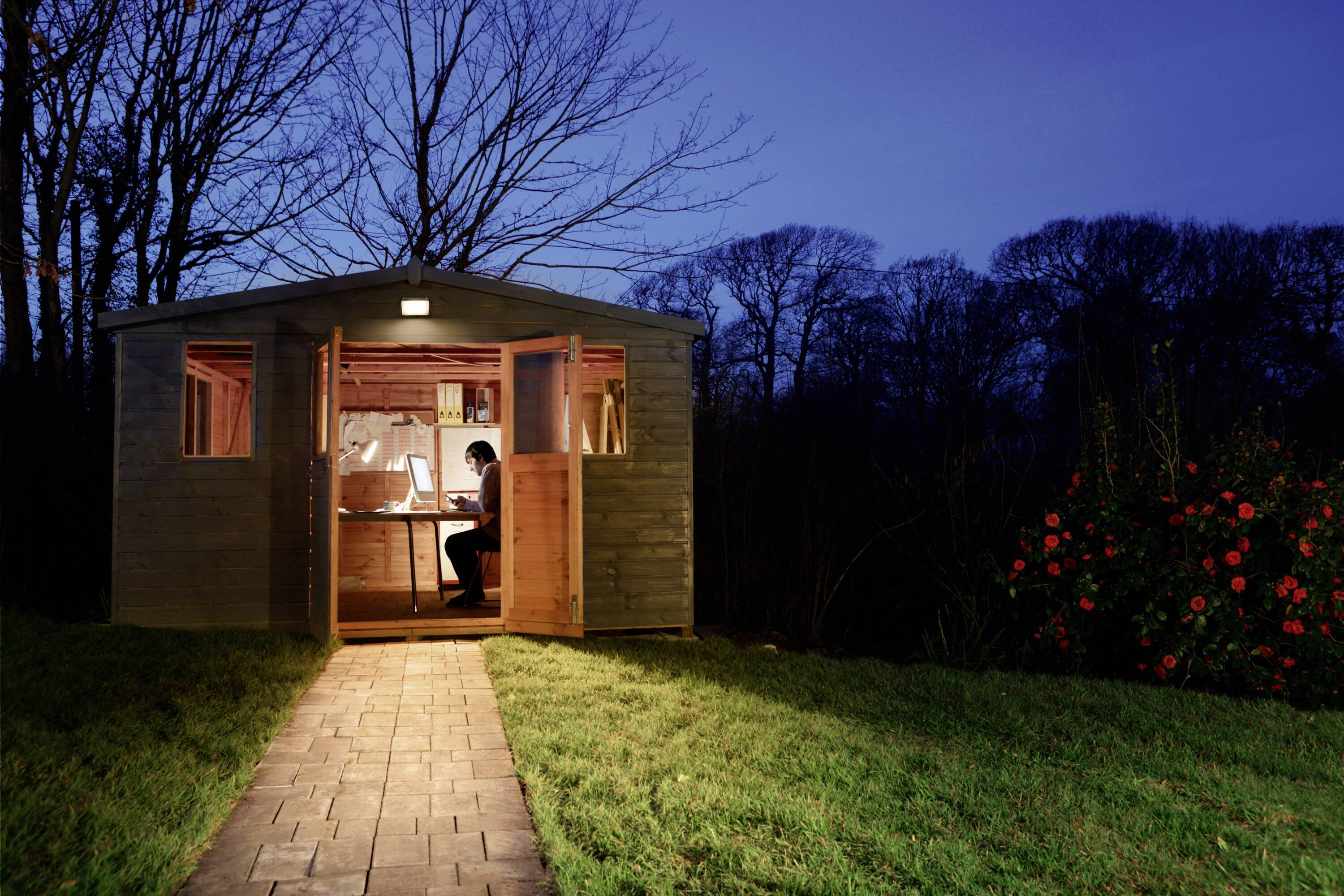 Eine Person arbeitet abends in einem beleuchteten Gartenhaus, umgeben von Bäumen und einem blühenden Busch.