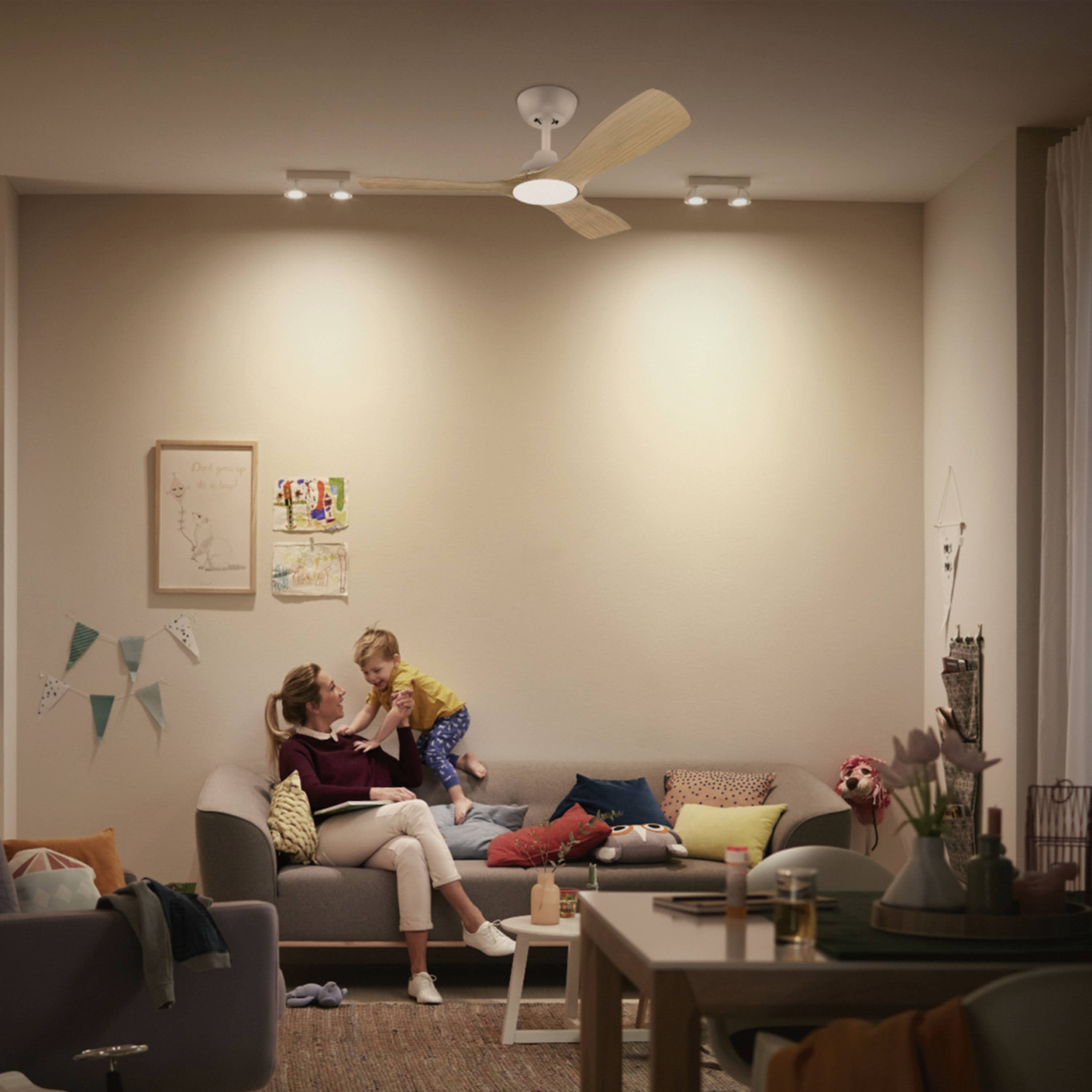 Eine Frau sitzt mit einem Kind auf einem Sofa in einem gemütlich beleuchteten Wohnzimmer, dekoriert mit Wandbildern und bunten Kissen.