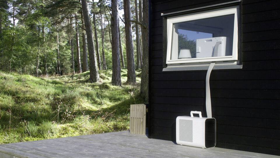 Ein tragbares Klimagerät steht auf einer Holzterrasse vor einem schwarzen Haus mit bewaldetem Hintergrund. Ein Schlauch führt zum Fenster.