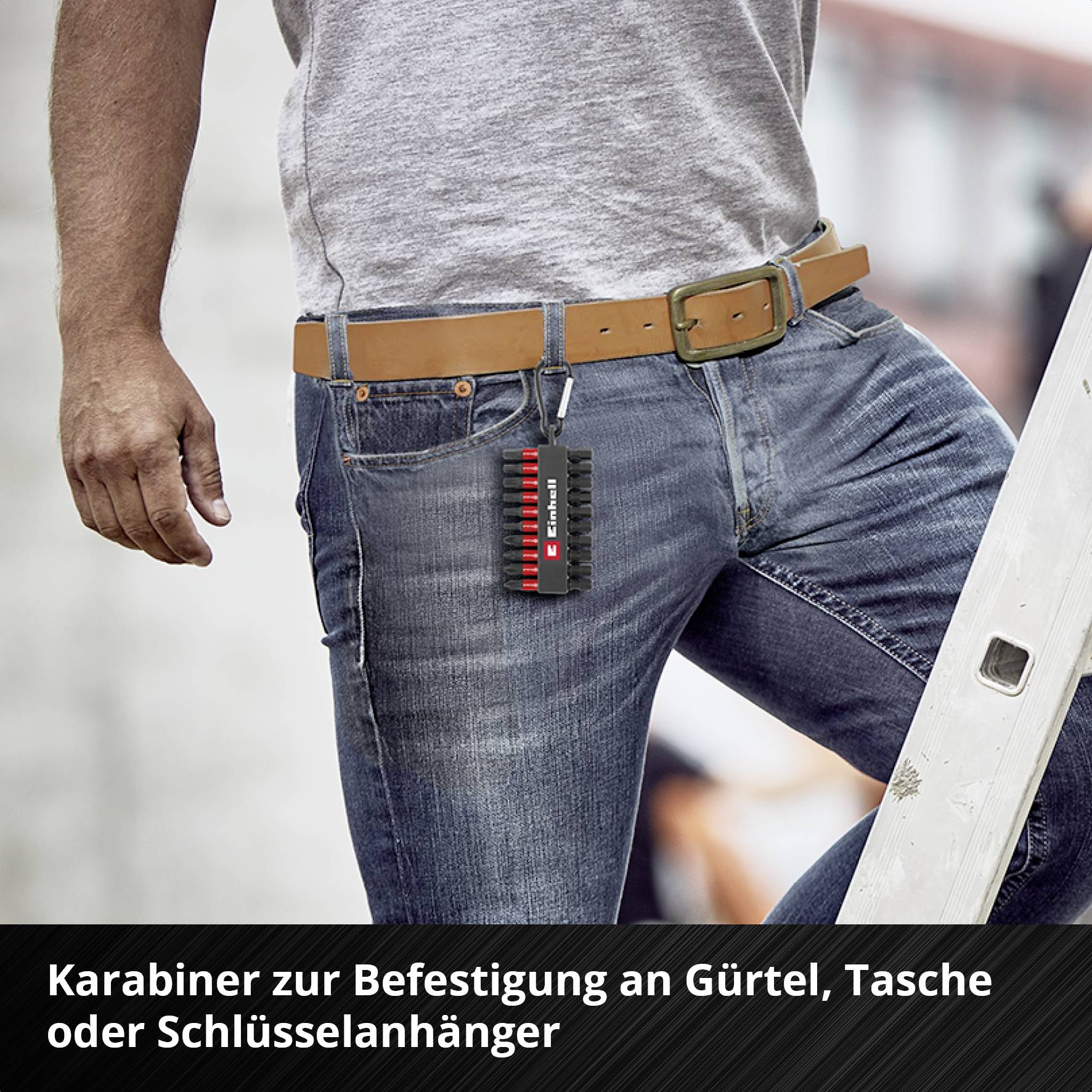 Mann mit Jeans befestigt einen Karabiner am Gürtel, um eine Tasche oder Schlüssel anzuhängen. Text unten: 'Karabiner zur Befestigung an Gürtel, Tasche oder Schlüsselanhänger'.