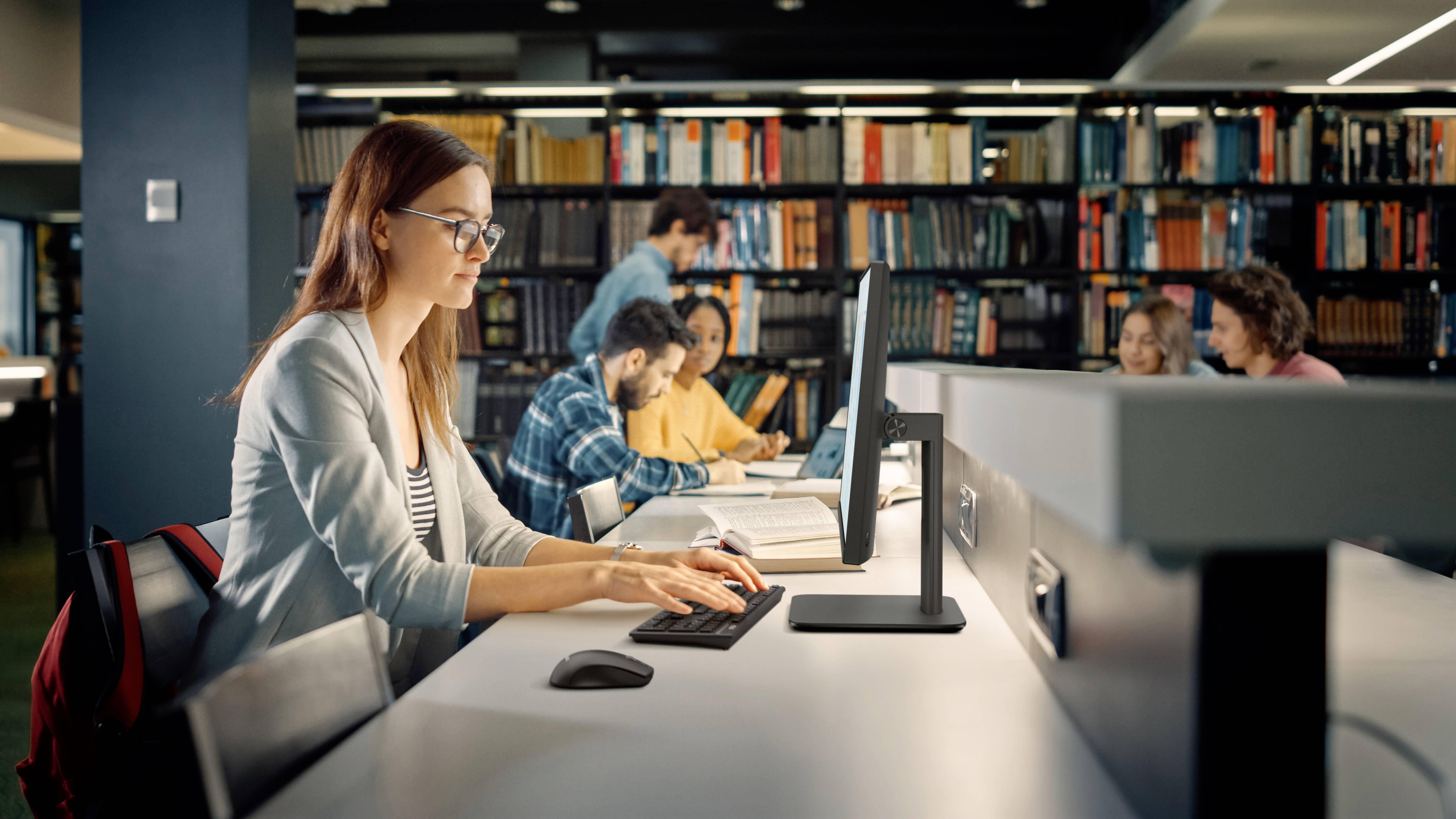 Eine Frau mit Brille arbeitet an einem Computer in einer Bibliothek. Mehrere Personen studieren im Hintergrund, umgeben von Regalen voller Bücher.