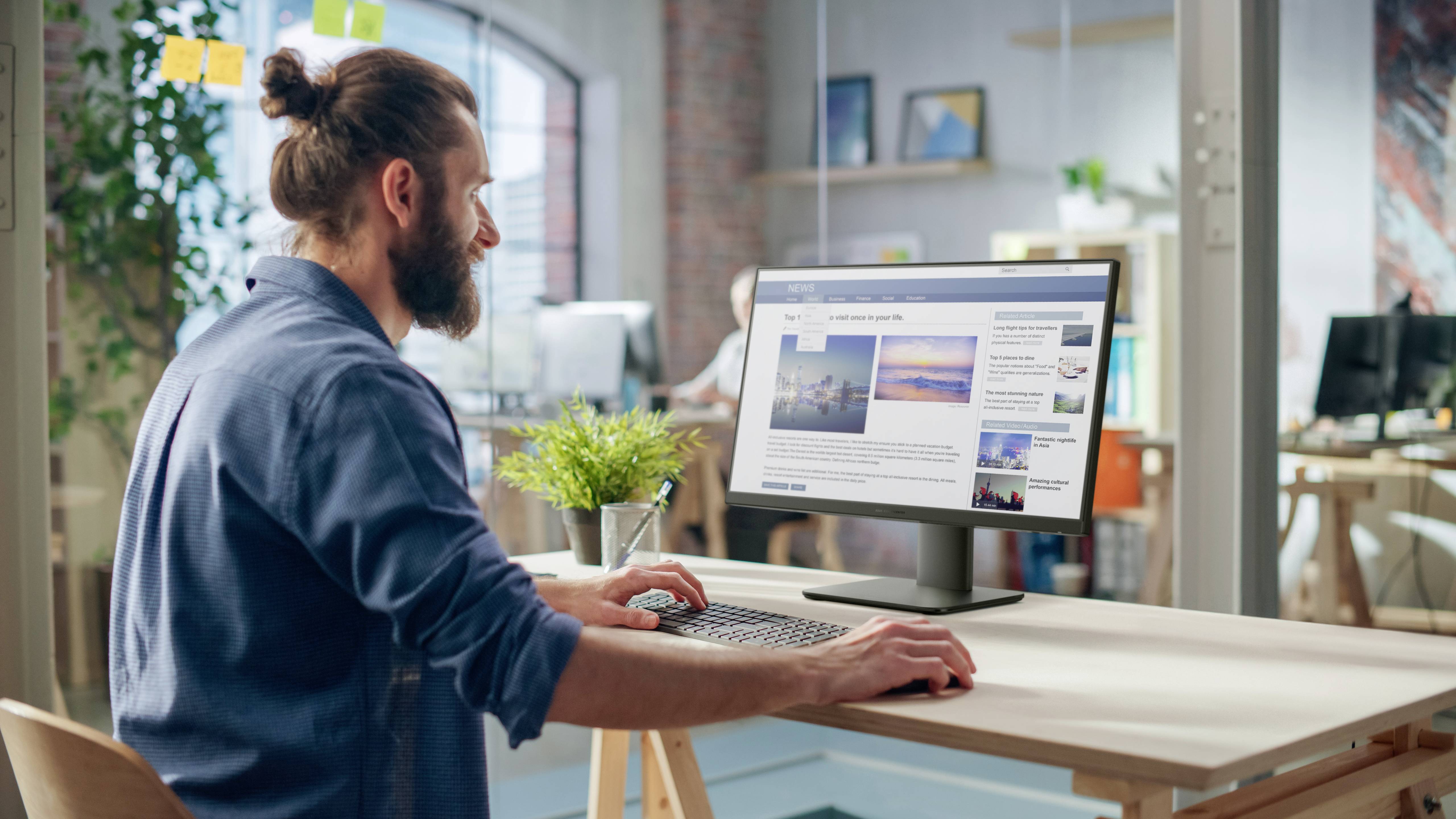 Eine Person mit Bart, die an einem Schreibtisch sitzt, blickt auf einen Desktop-Monitor, der Bilder und Text in einem modernen Büroumfeld zeigt.