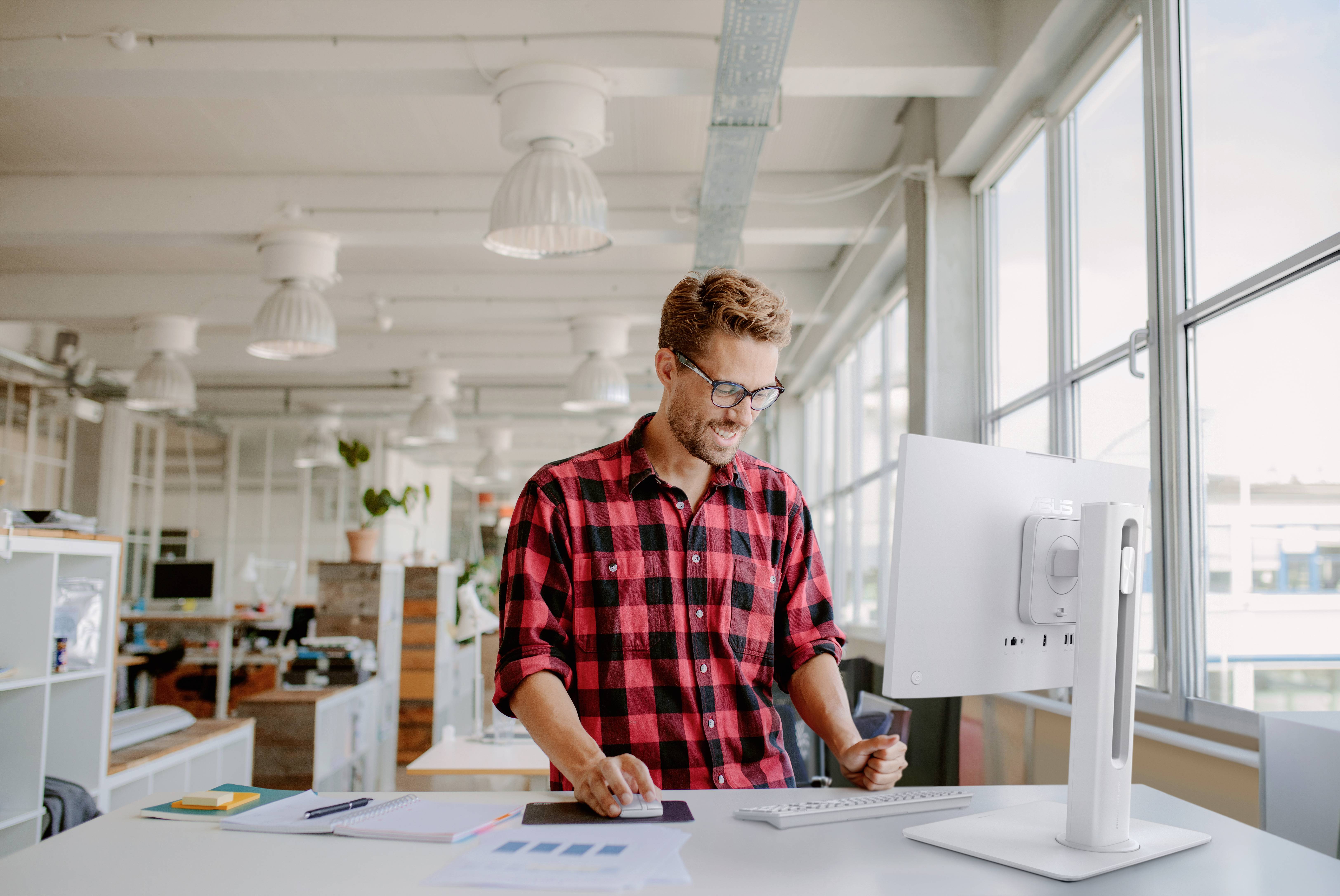 Eine Person in einem roten Karohemd steht lächelnd in einem hellen Büro und arbeitet an einem Computer. Natürliches Licht strömt durch große Fenster.