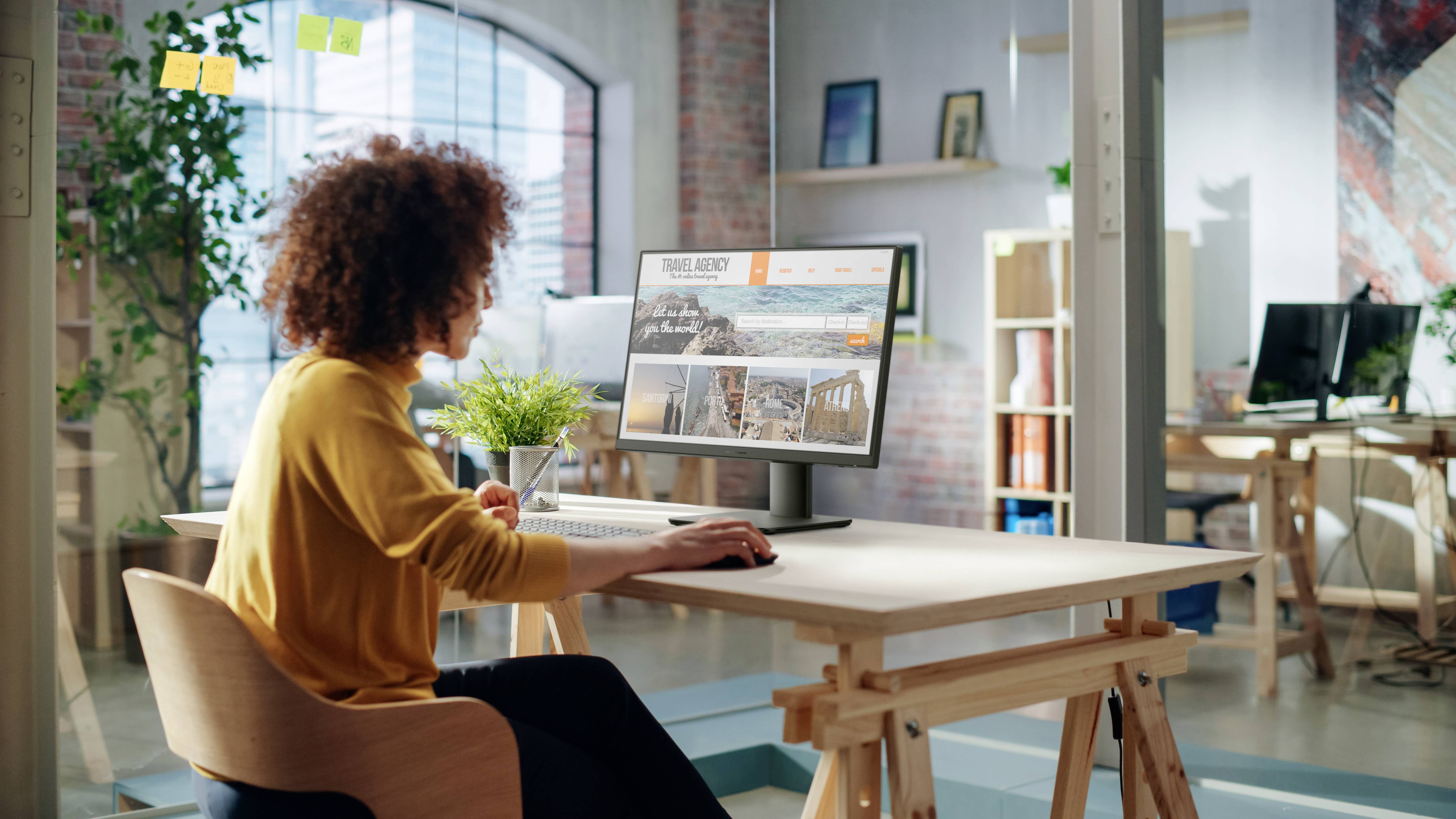 Eine Person sitzt an einem Schreibtisch und verwendet einen Computer, auf dem eine Immobilienwebseite geöffnet ist. Der Raum verfügt über große Fenster, Pflanzen und Regale.