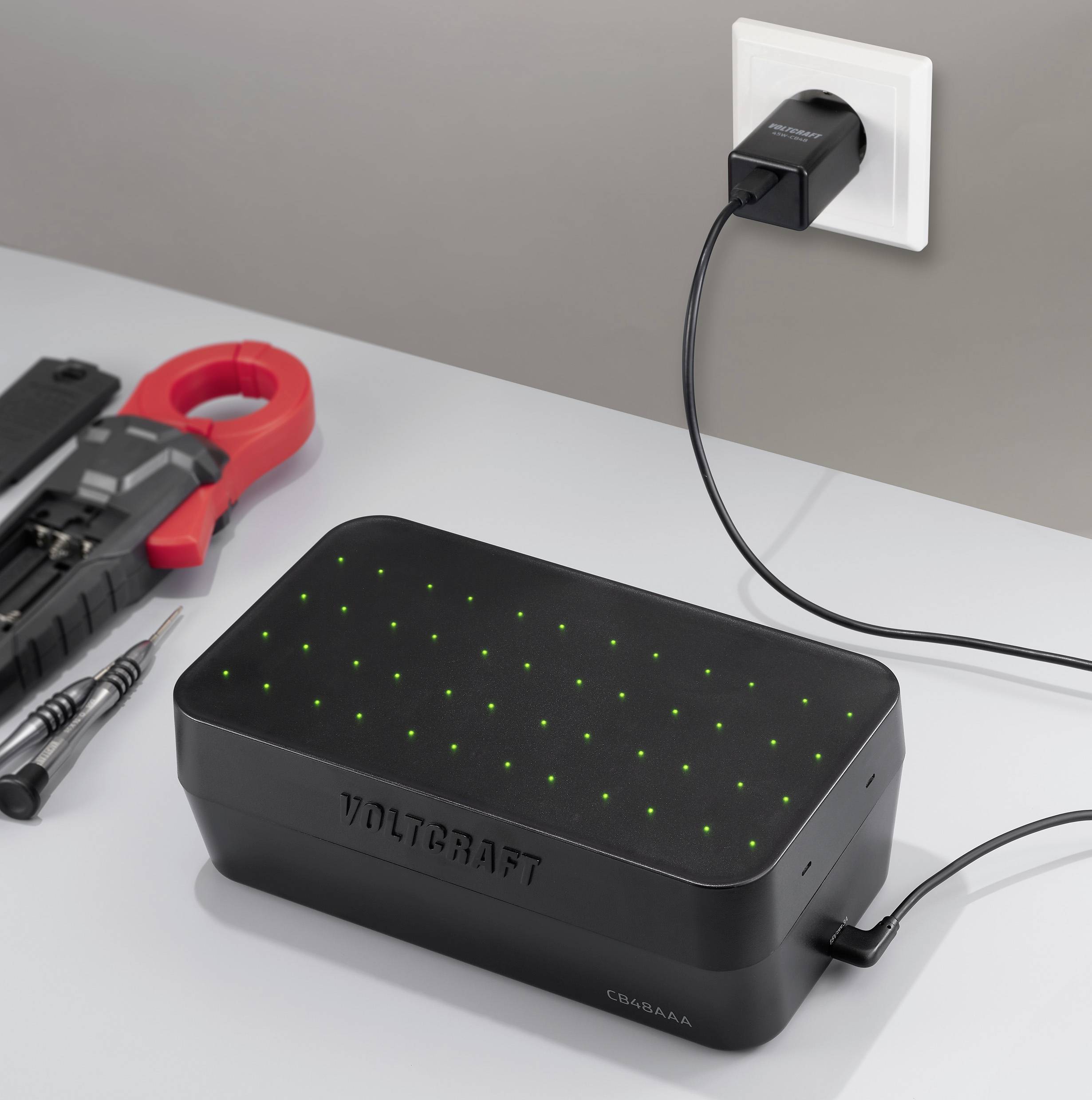 Ein 'Voltcraft' Ladegerät auf einem Tisch mit Werkzeugen in der Nähe, in eine Steckdose eingesteckt. Lichter zeigen den Ladestatus an.