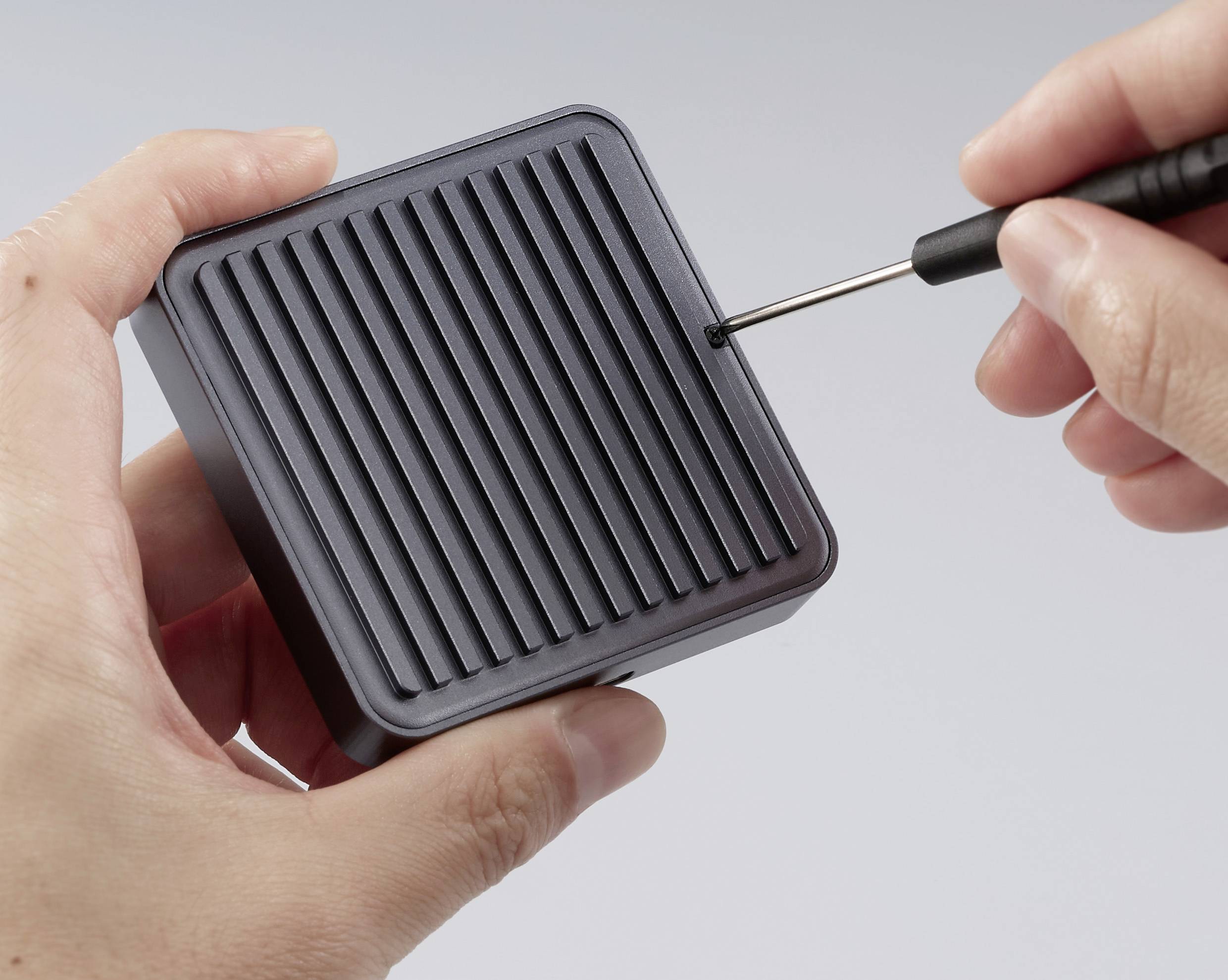 Eine Person benutzt einen Schraubendreher, um ein schwarzes, quadratisches elektronisches Gerät mit gerillter Oberfläche zu öffnen, was auf Wartung oder Reparatur hindeutet.