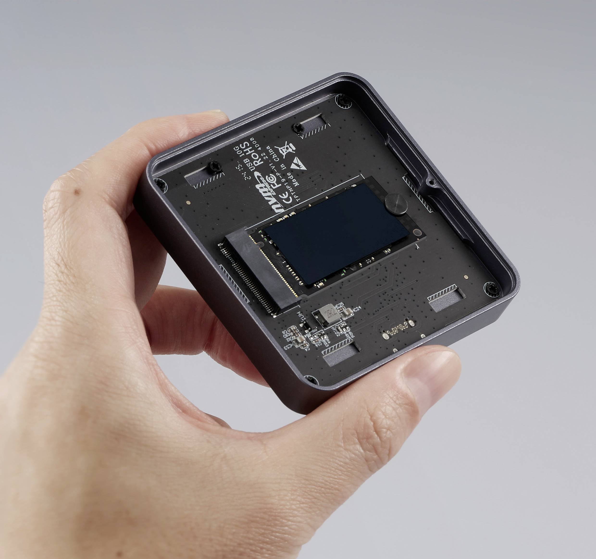 Eine Hand, die ein kompaktes elektronisches Gerät mit freiliegendem Leiterplattencircuit hält, mit einem zentralen schwarzen rechteckigen Chip vor einem grauen Hintergrund.