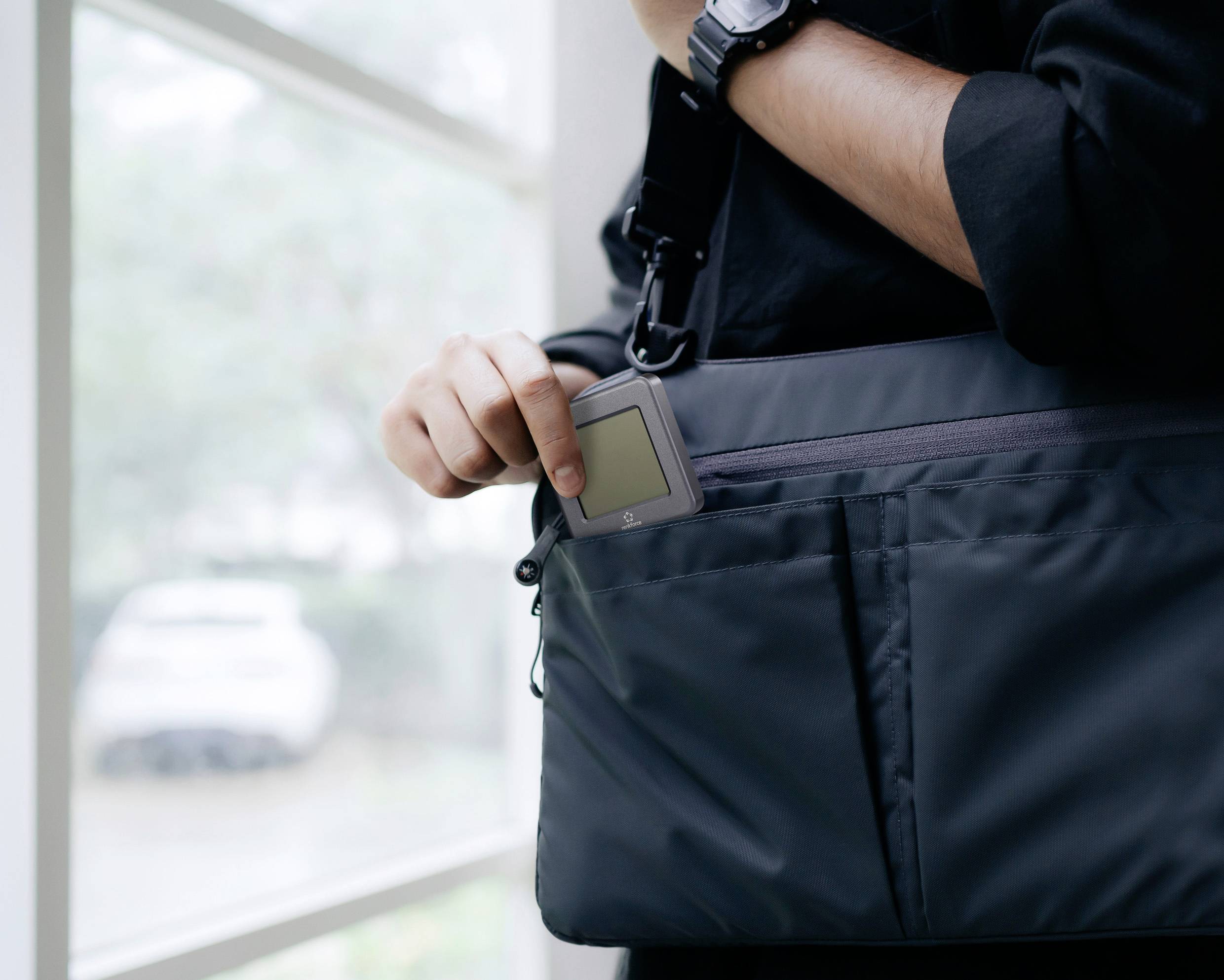 Eine Person hält ein schlankes elektronisches Gerät, während sie es in die Vordertasche einer schwarzen Umhängetasche in der Nähe eines Fensters legt.