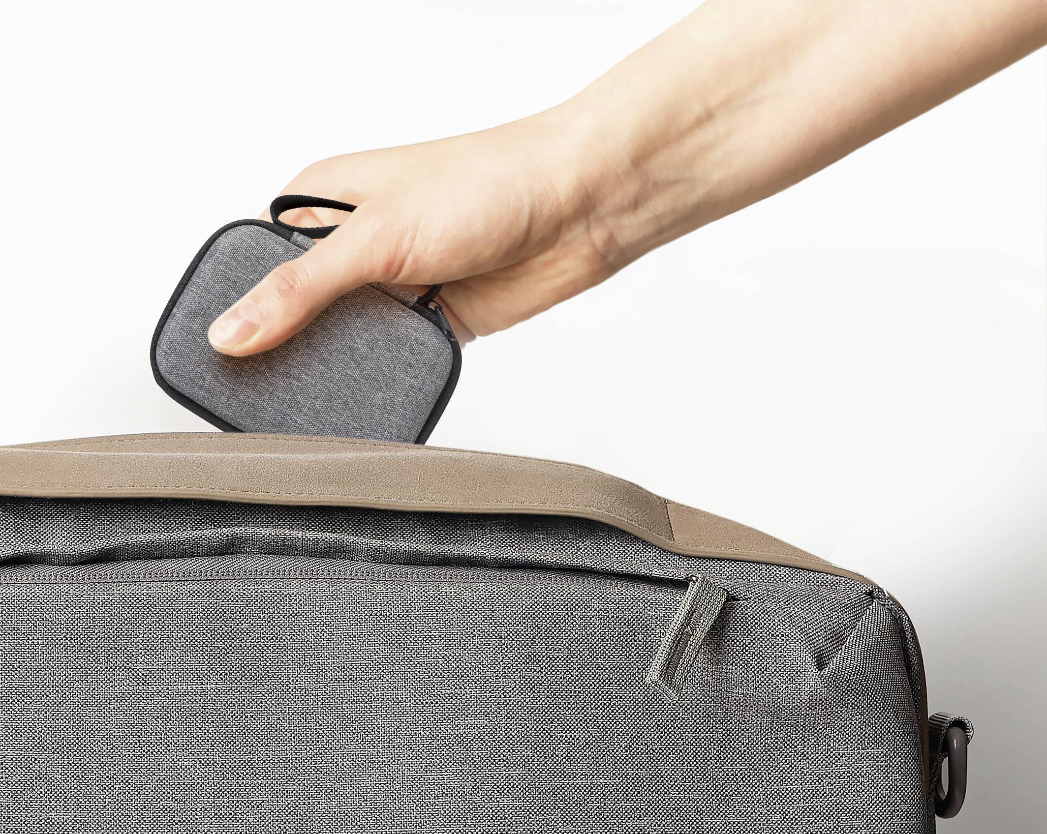 Eine Hand legt einen kleinen, grauen, mit Stoff bezogenen Gegenstand in die Reißverschlusstasche einer grauen und braunen Tasche, was auf Reise oder Aufbewahrung hindeutet.