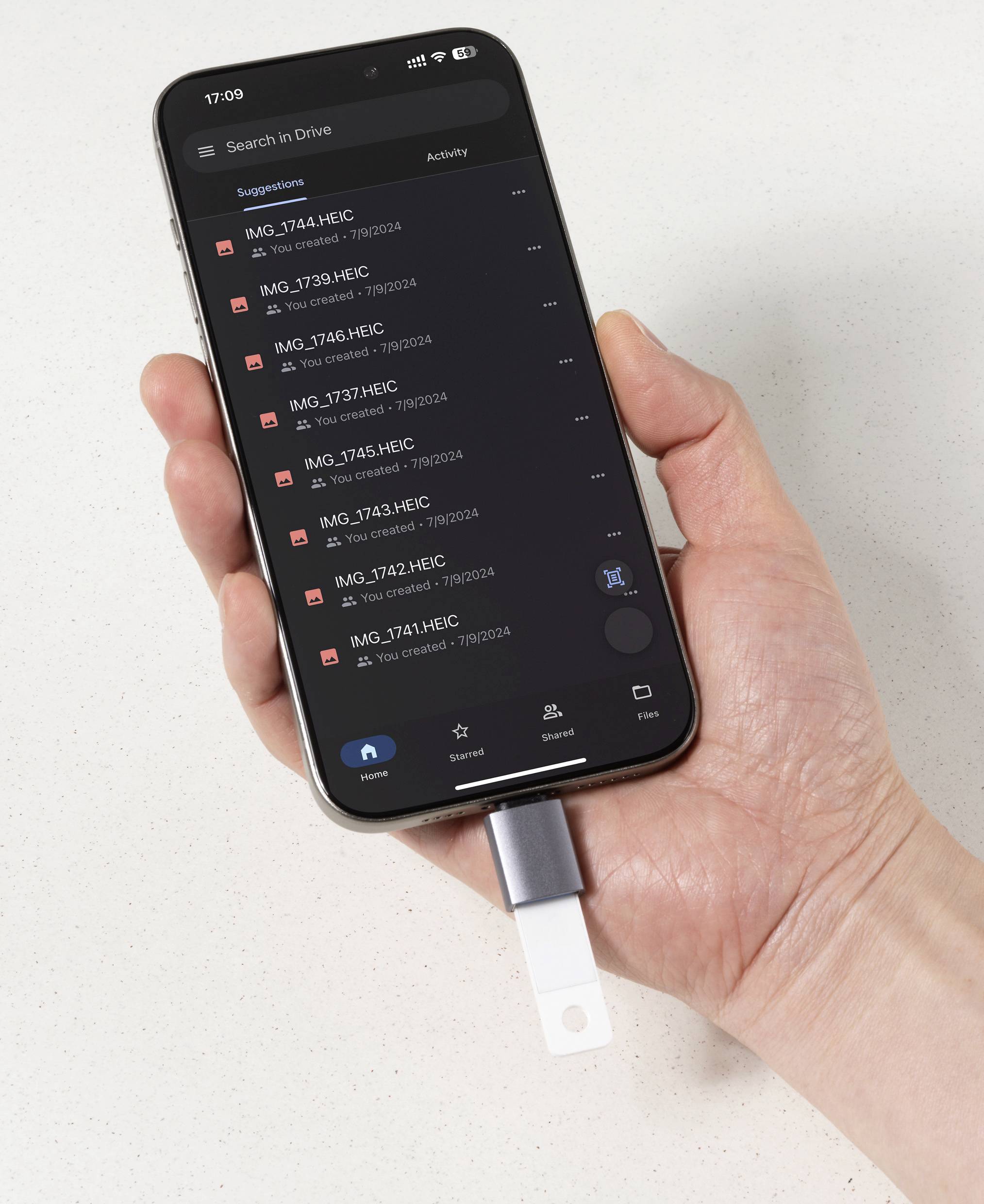 Eine Hand, die ein Smartphone hält, das eine Liste von Bilddateien in einer Cloud-Speicher-App anzeigt. Ein USB-Stick ist an das Telefon angeschlossen.