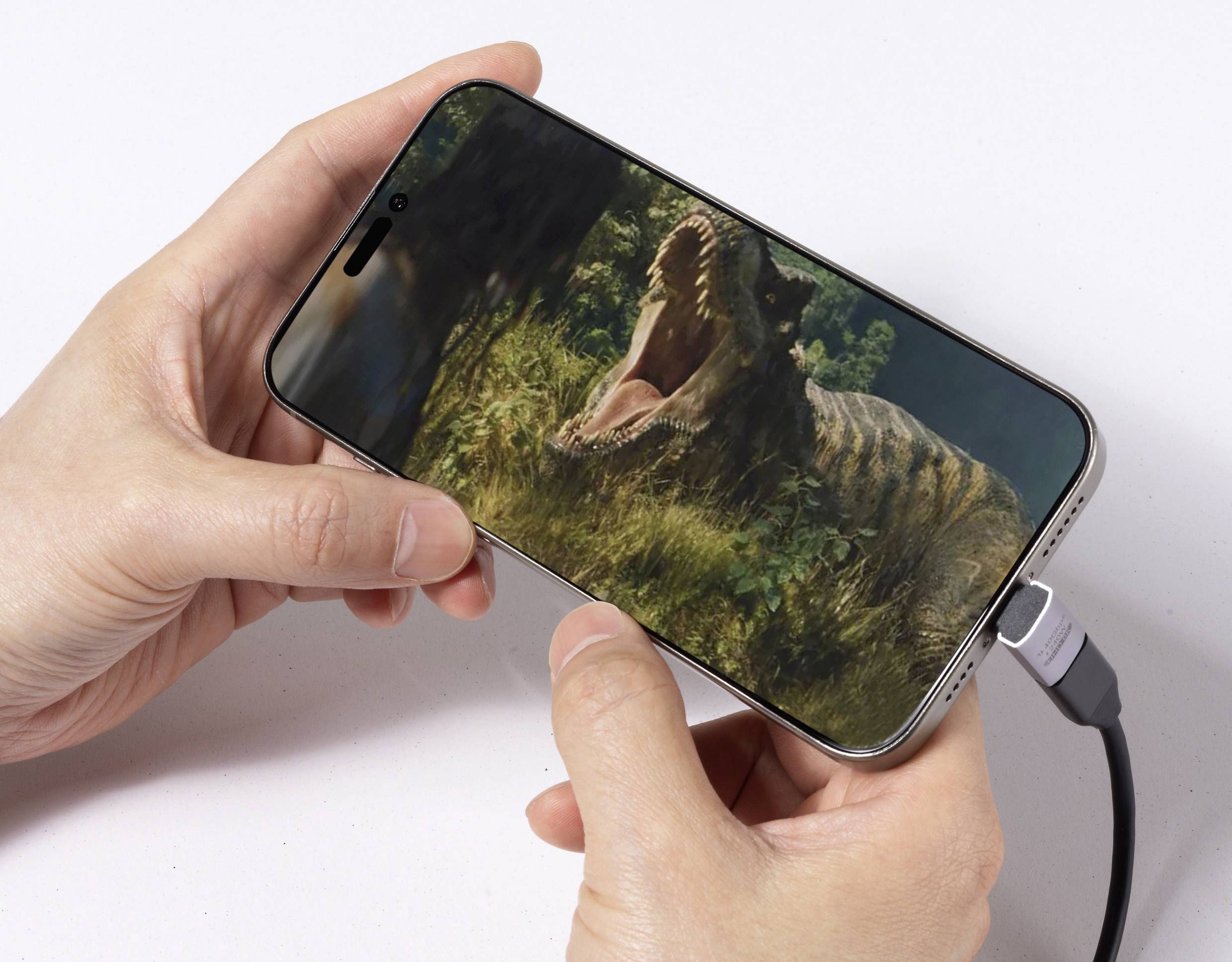 Eine Person, die ein Smartphone hält, das eine Szene eines Dinosauriers zeigt, der in dichtem Blattwerk brüllt, wobei das Telefon mit einem Ladekabel verbunden ist.