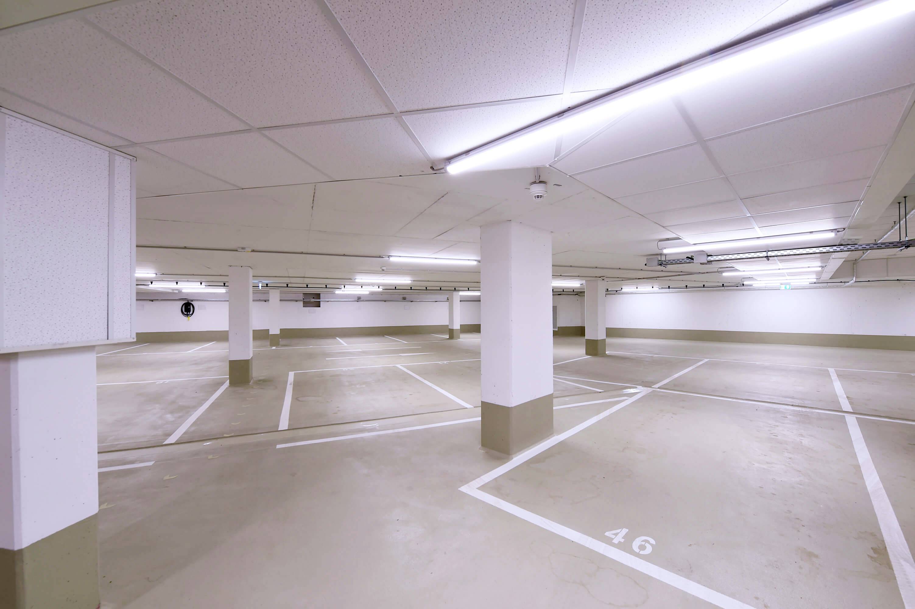 Parkhaus mit leeren Stellplätzen, weißer Decke und markierten Parklinien. Deckenleuchten erhellen den Raum, eine Kamerasicht erscheint links.