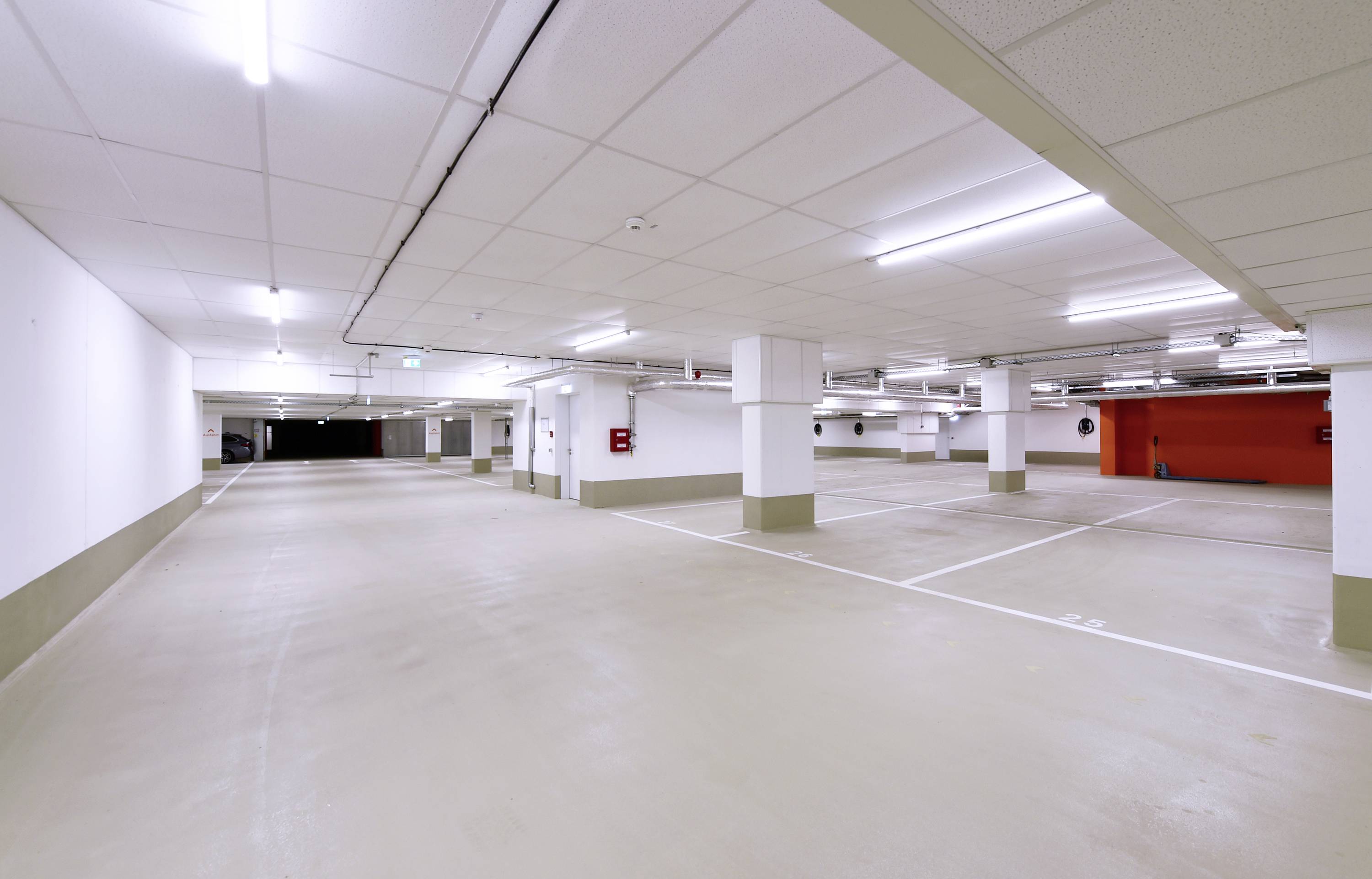 Leerer Tiefgaragen-Parkplatz mit weißen Markierungen und Leuchtstoffbeleuchtung. Wände sind teils rot und teils weiß gestrichen.