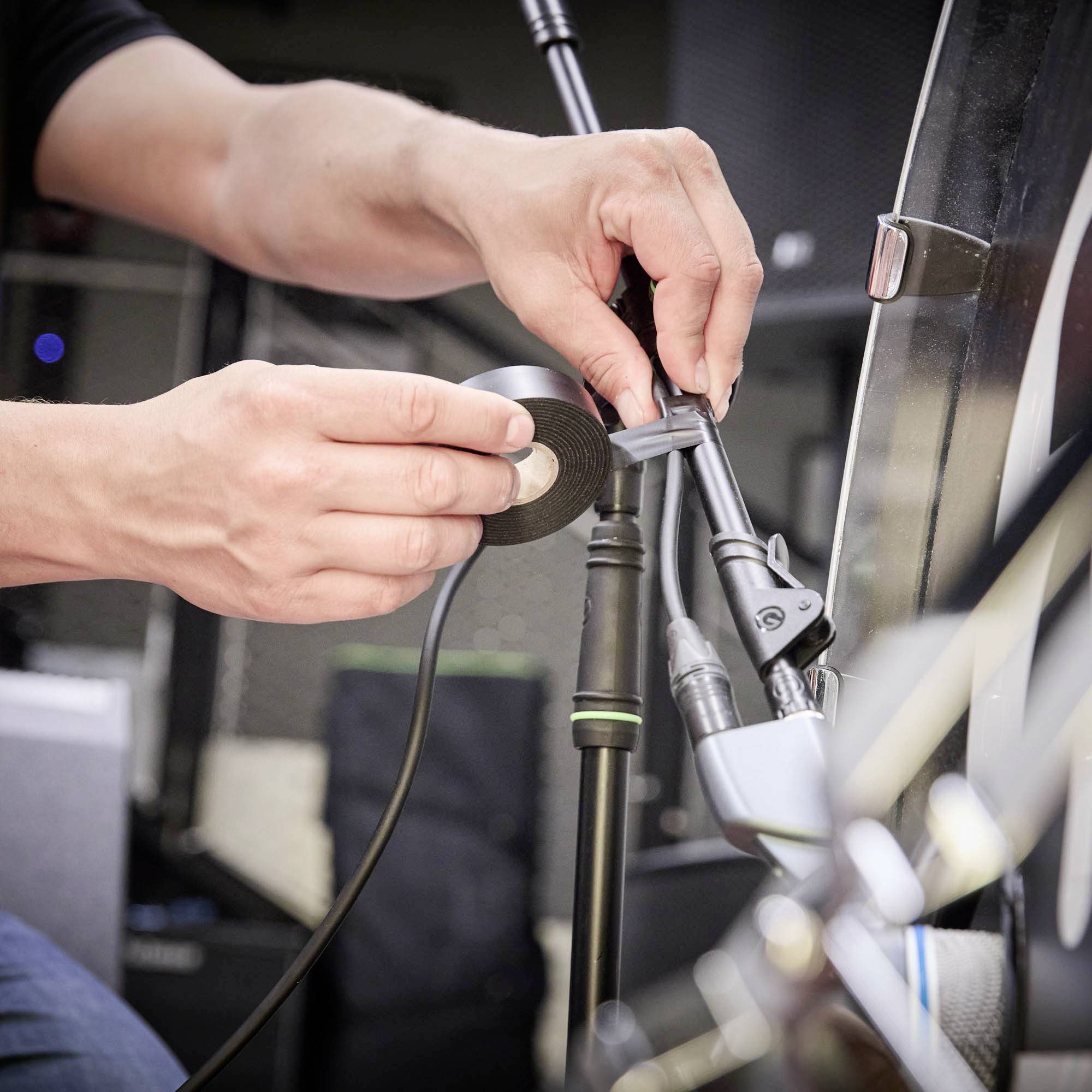 Eine Person befestigt ein Mikrofon an einem Ständer, hält Kabel und Klebeband. Aufnahme im Innenraum mit technischer Ausrüstung.