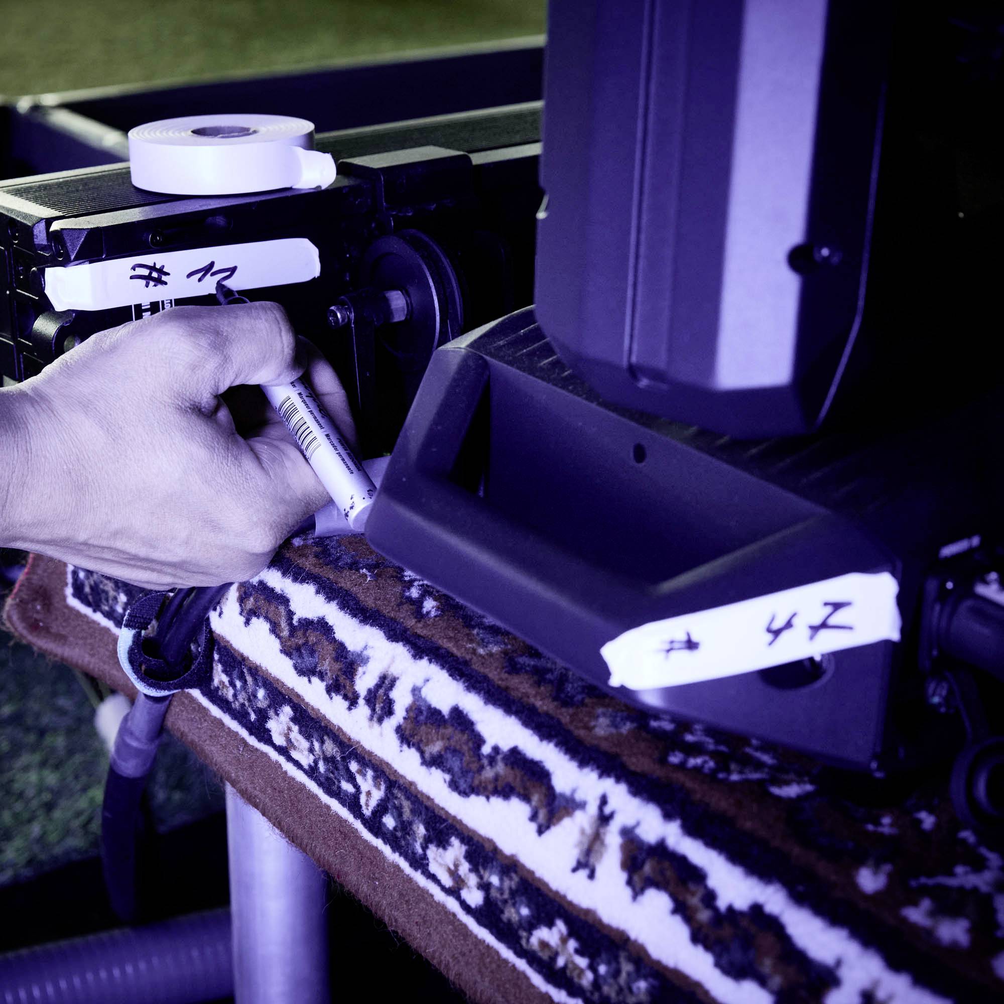 Eine Hand hält einen schwarzen Marker und beschriftet ein schwarzes Gerät. Auf dem Gerät sind handgeschriebene Etiketten sichtbar.