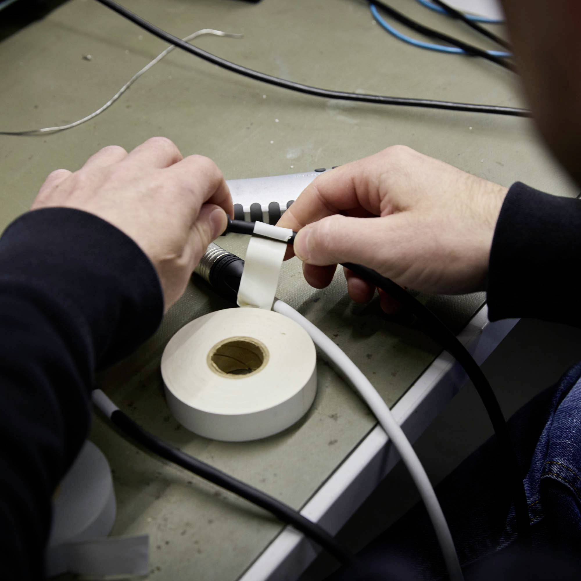 Eine Person klebt ein weißes Band um ein Kabel auf einem Tisch. Daneben liegt eine Rolle mit ähnlichem Band.