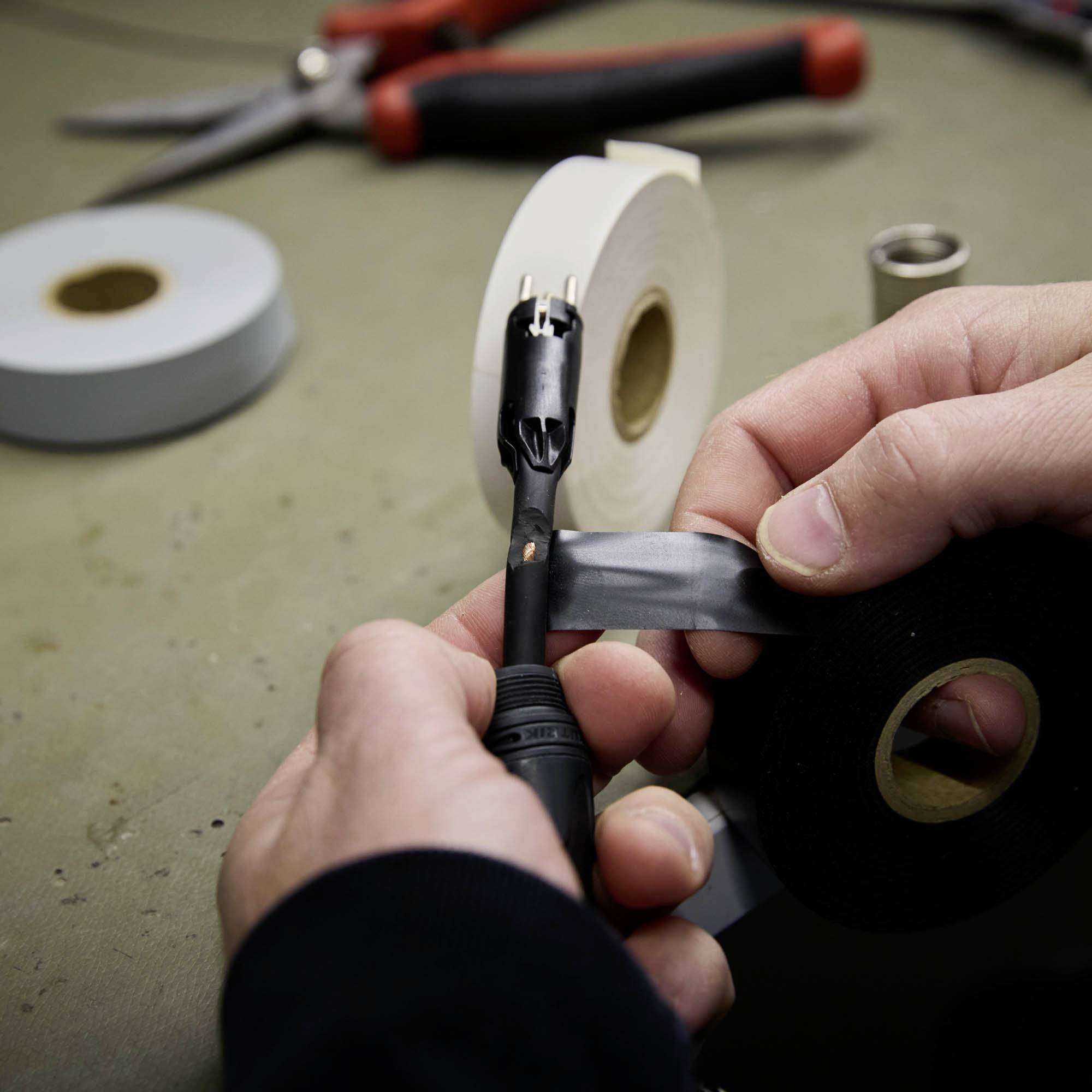 Nahaufnahme von Händen, die ein Kabel mit schwarzem Isolierband umwickeln. Im Hintergrund liegen Scheren und Klebebandrollen.