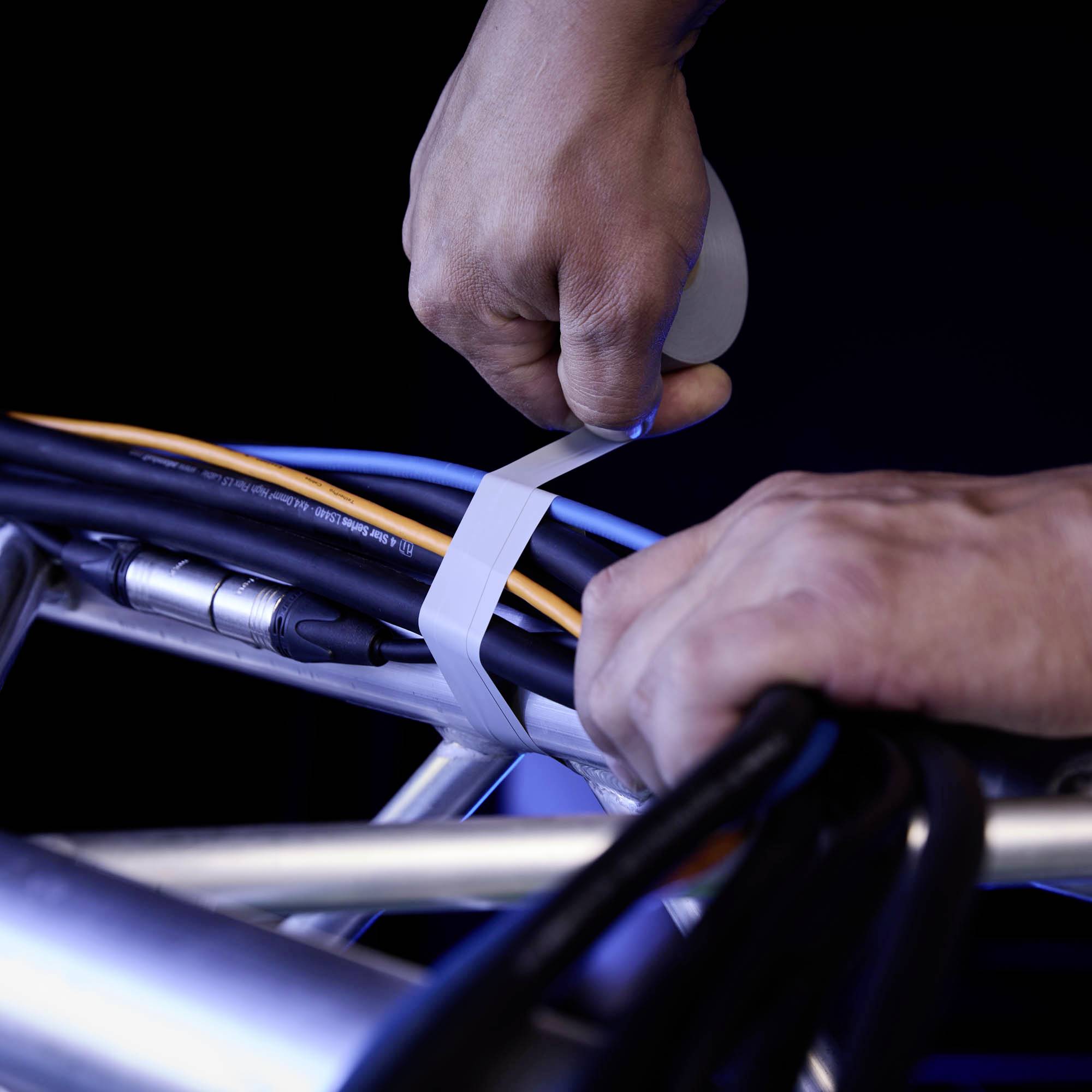 Zwei Hände binden Kabel mit einem Kabelbinder an ein silbernes Gestell. Fokus auf Kabelmanagement, Technik im Hintergrund.