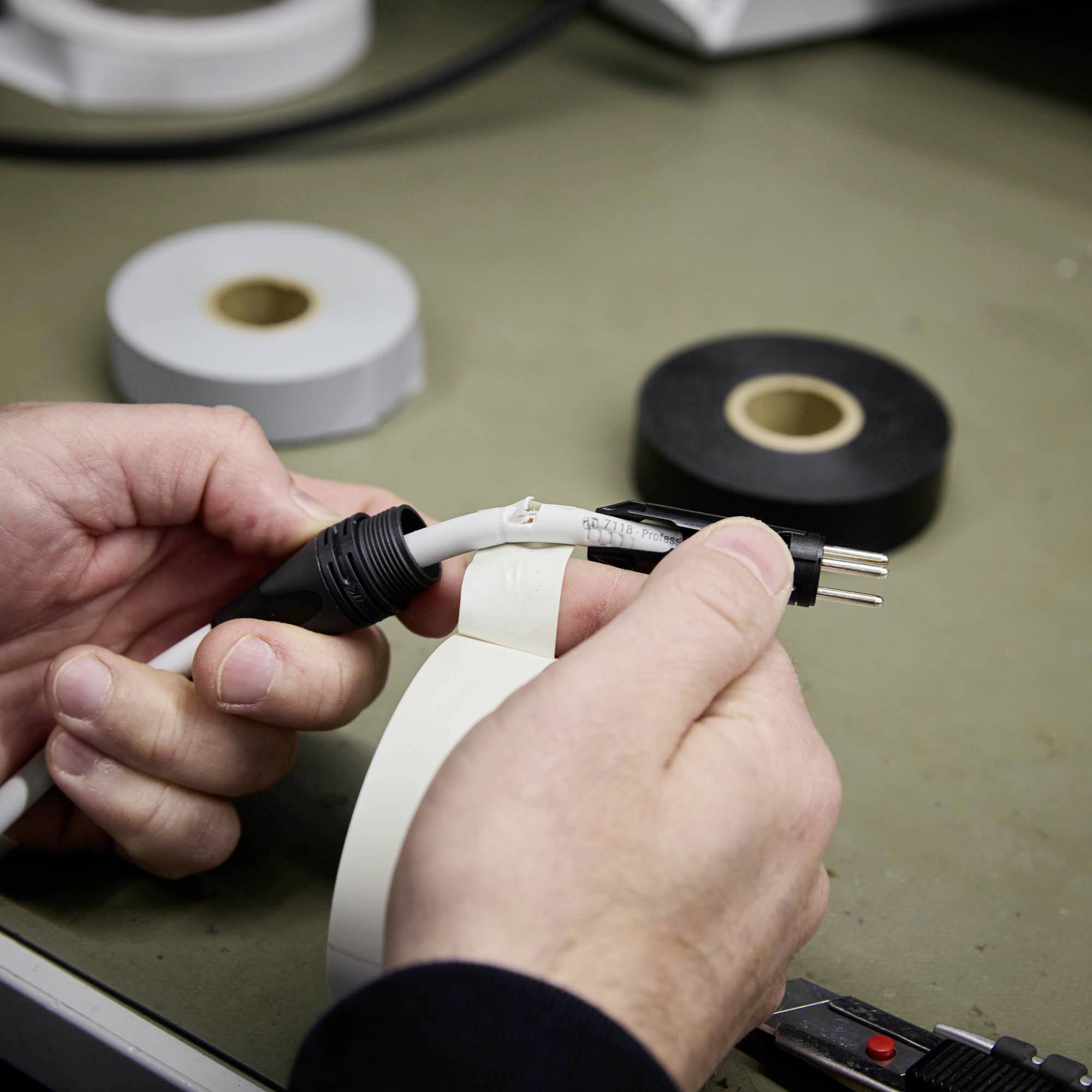 Eine Person repariert ein Stromkabel mit Isolierband. Im Hintergrund sind zwei Rollen Klebeband zu sehen, eine weiße und eine schwarze.