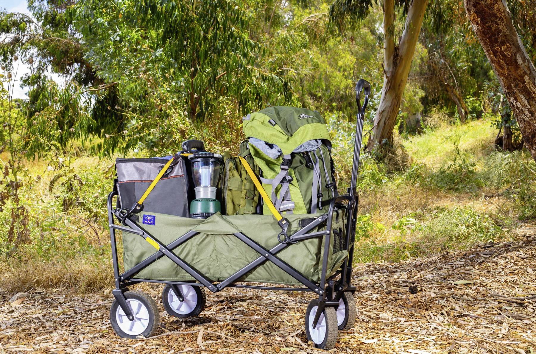 Ein Faltwagen ist mit Campingausrüstung gefüllt, darunter ein Rucksack, eine Laterne und eine Kühltasche, und steht in einem bewaldeten Gebiet mit Bäumen im Hintergrund.