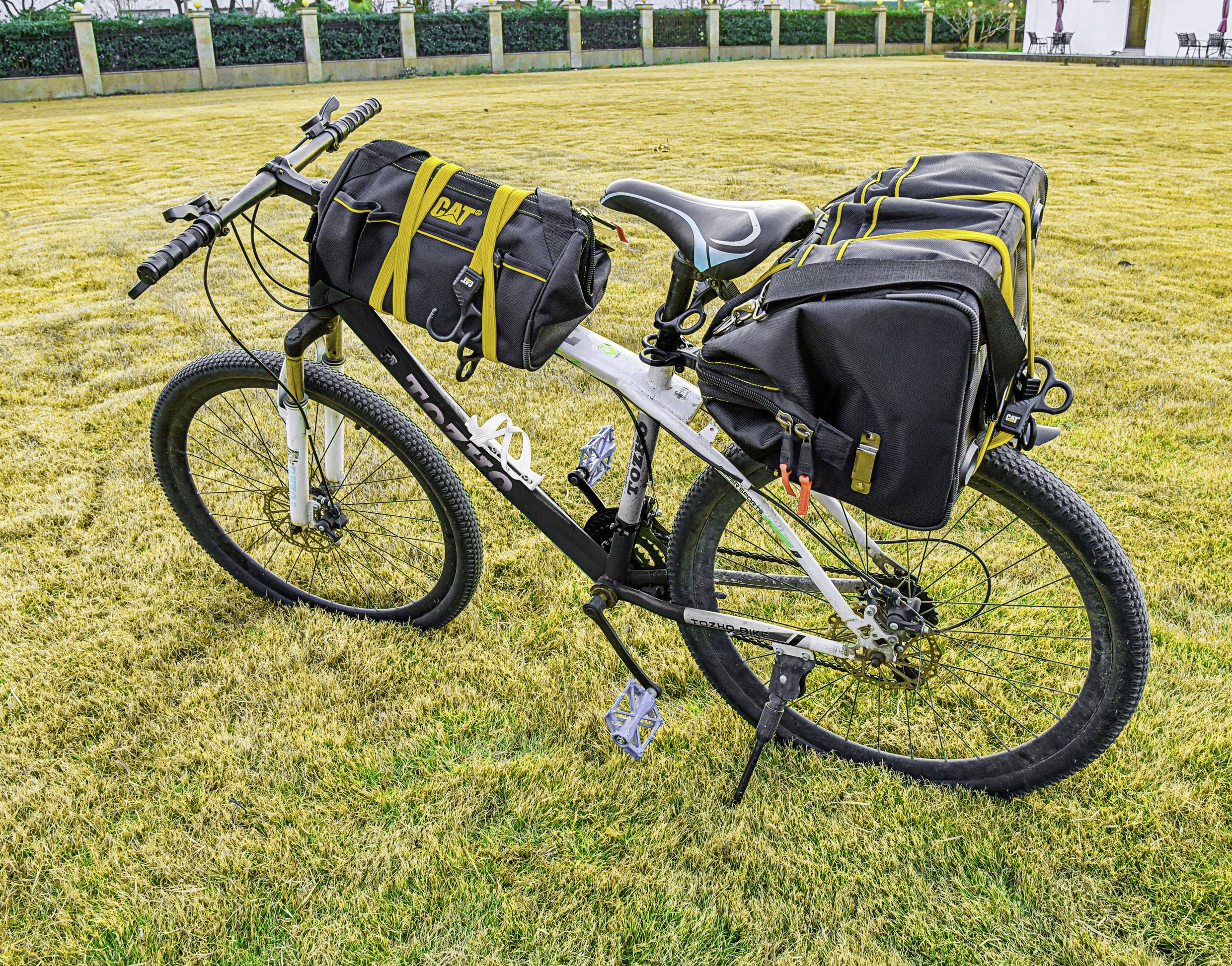 Ein Fahrrad mit großen, schwarzen Satteltaschen und einer Lenkertasche, geparkt auf trockenem Gras in einem Park mit einem Zaun im Hintergrund.
