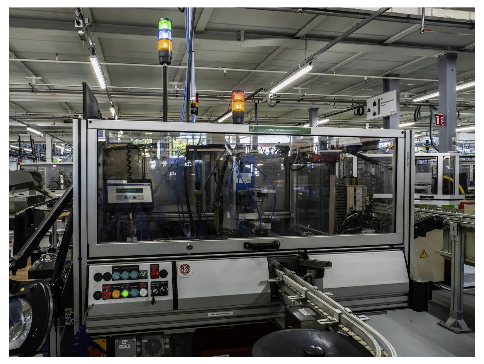 Automatisierte Industriemaschine auf einem Fabrikboden, umschlossen von Sicherheitsglas mit Statusleuchten und Steuertasten, Material auf einem Förderband handhabend.