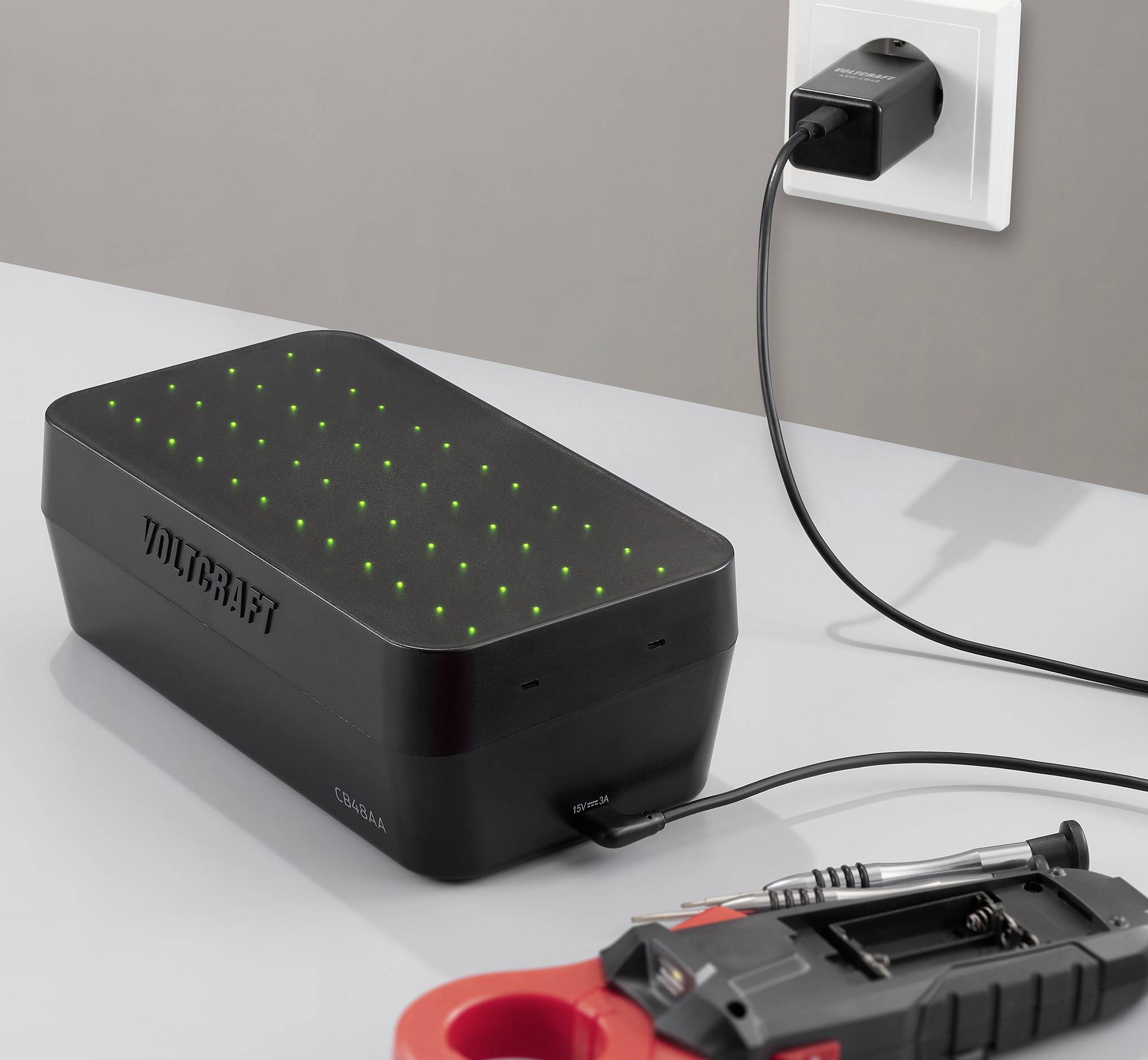 Ein schwarzes Gerät mit grünen LED-Leuchten ist in eine Wandsteckdose eingesteckt, neben einem roten Werkzeugkoffer mit einem Schraubendreher, auf einer grauen Oberfläche.