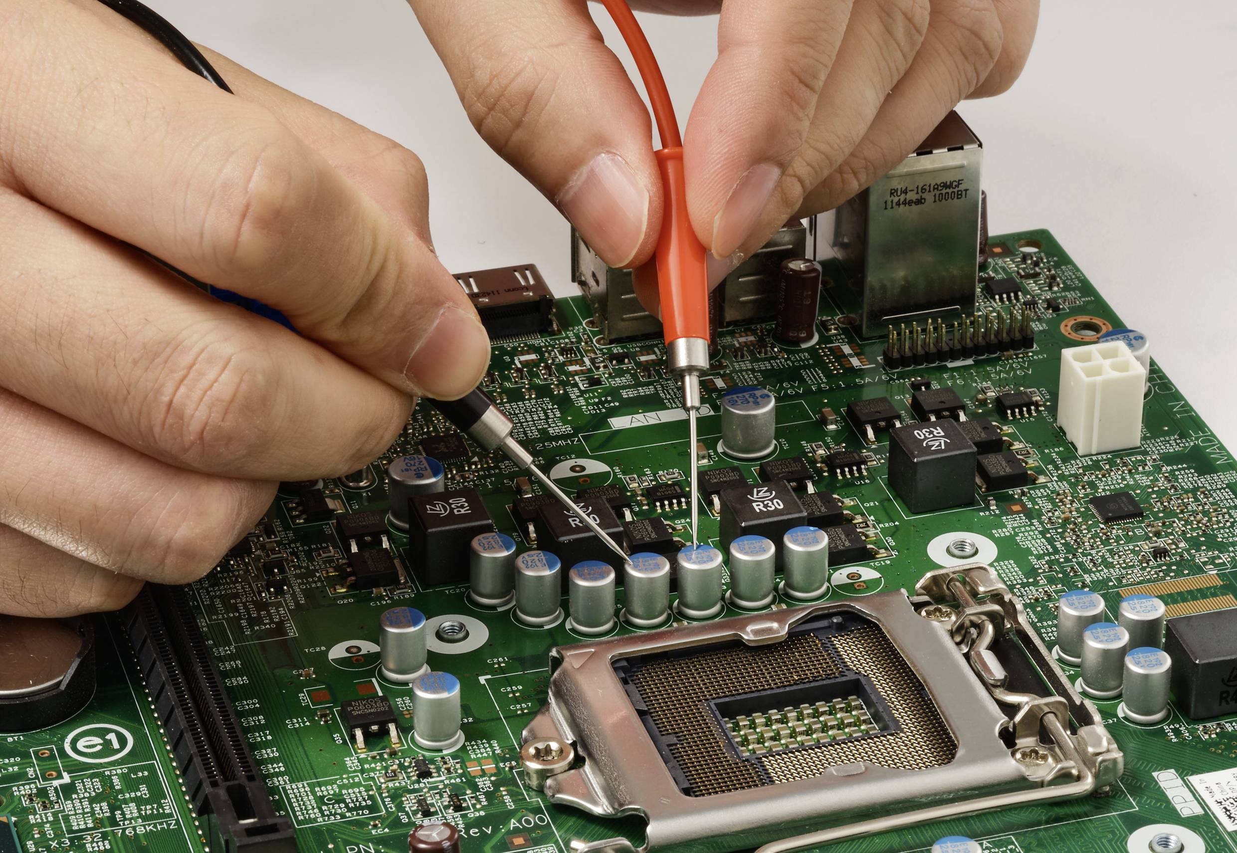 Nahaufnahme von Händen, die Multimeter-Messspitzen verwenden, um eine Leiterplatte zu testen. Die Leiterplatte zeigt verschiedene elektronische Komponenten.