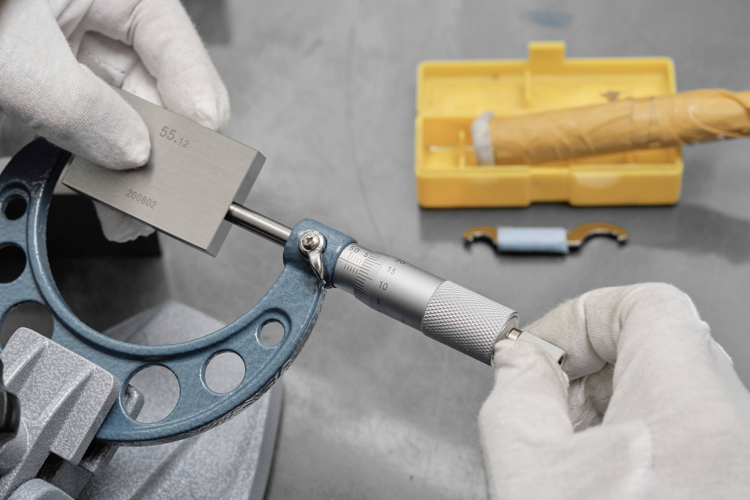 Eine Person misst einen Metallblock, der mit 'S5 1.2' beschriftet ist, mit einem Mikrometerschrauber. Werkzeuge liegen auf dem Tisch, darunter ein gelbes Etui.