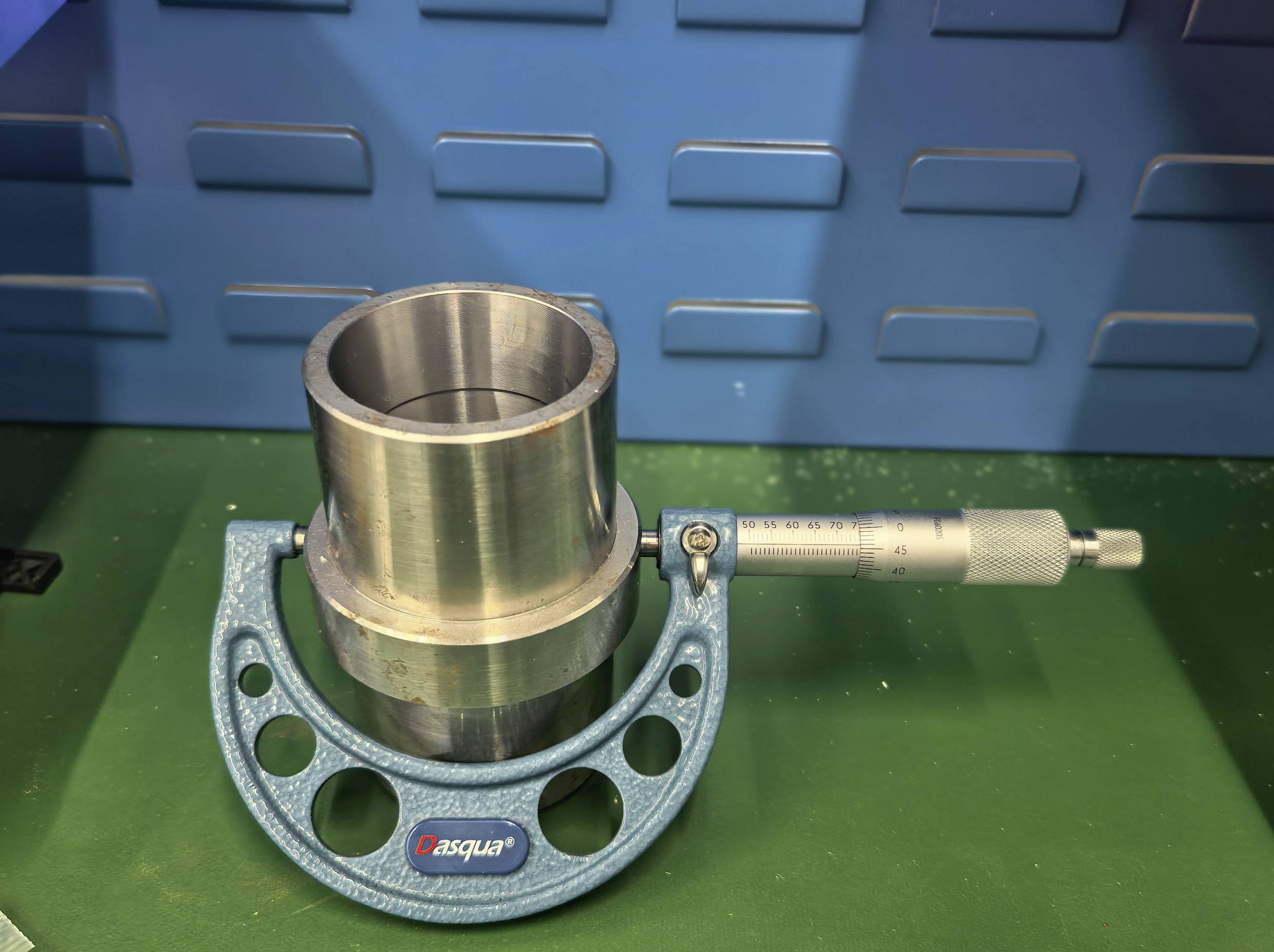 Ein Metallzylinder in einem Mikrometer, auf einer grünen Oberfläche mit einem blauen strukturierten Hintergrund, verwendet für präzise Messungen.