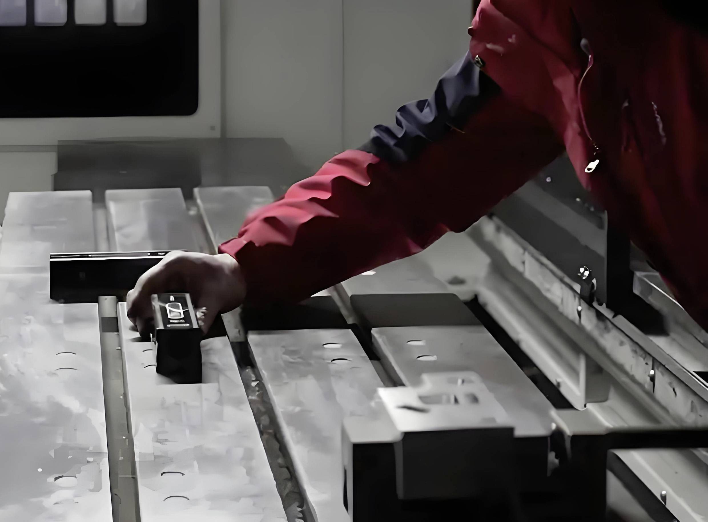 Eine Person in einer roten Jacke bedient eine Metallbearbeitungsmaschine und justiert eine Komponente auf einem Metalltisch in einer industriellen Umgebung.