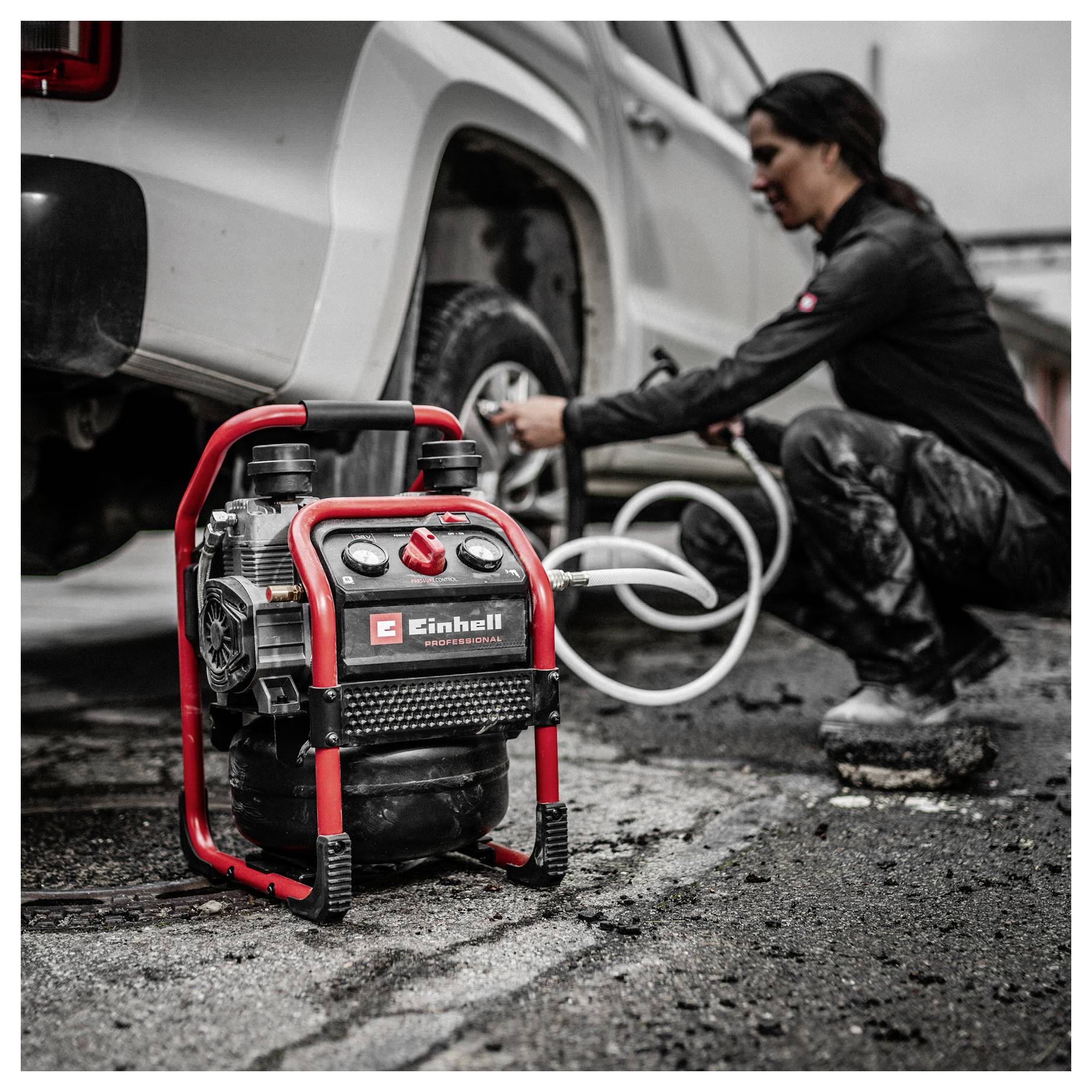Ein tragbarer Luftkompressor im Vordergrund, während eine Frau ihn nutzt, um einen Reifen neben einem weißen Pickup-Truck auf nasser Fahrbahn aufzupumpen.