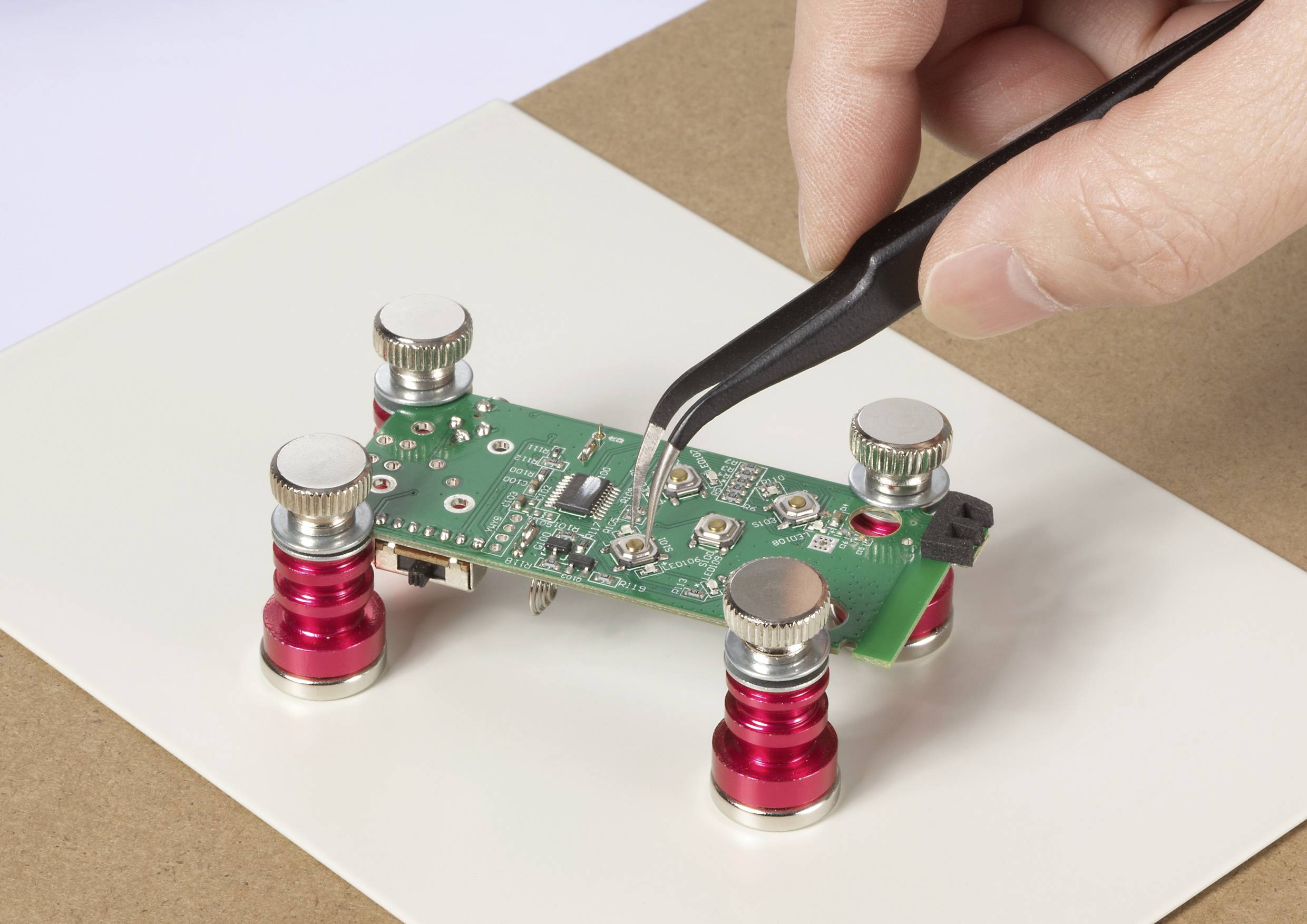 Eine Hand verwendet eine Pinzette, um eine Komponente auf eine grüne Leiterplatte zu platzieren, die von vier roten und silbernen verstellbaren Ständern gestützt wird.