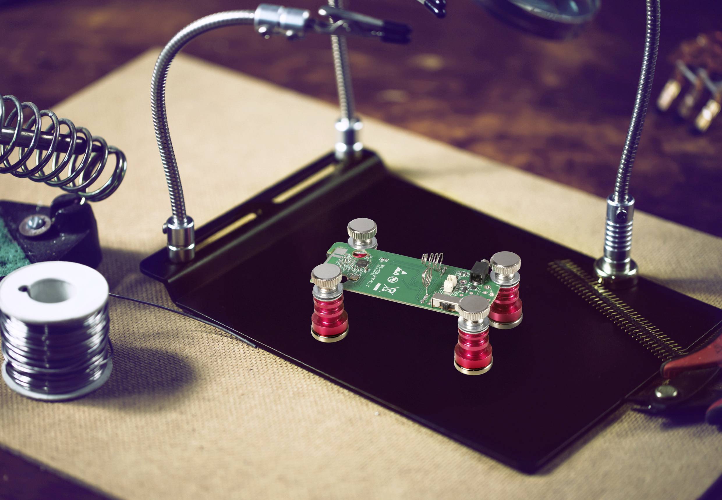 Ein kleines elektronisches Leiterplättchen ist auf einer Plattform mit vier roten Spulenstützen montiert, umgeben von einer Lupe und Werkzeugen.