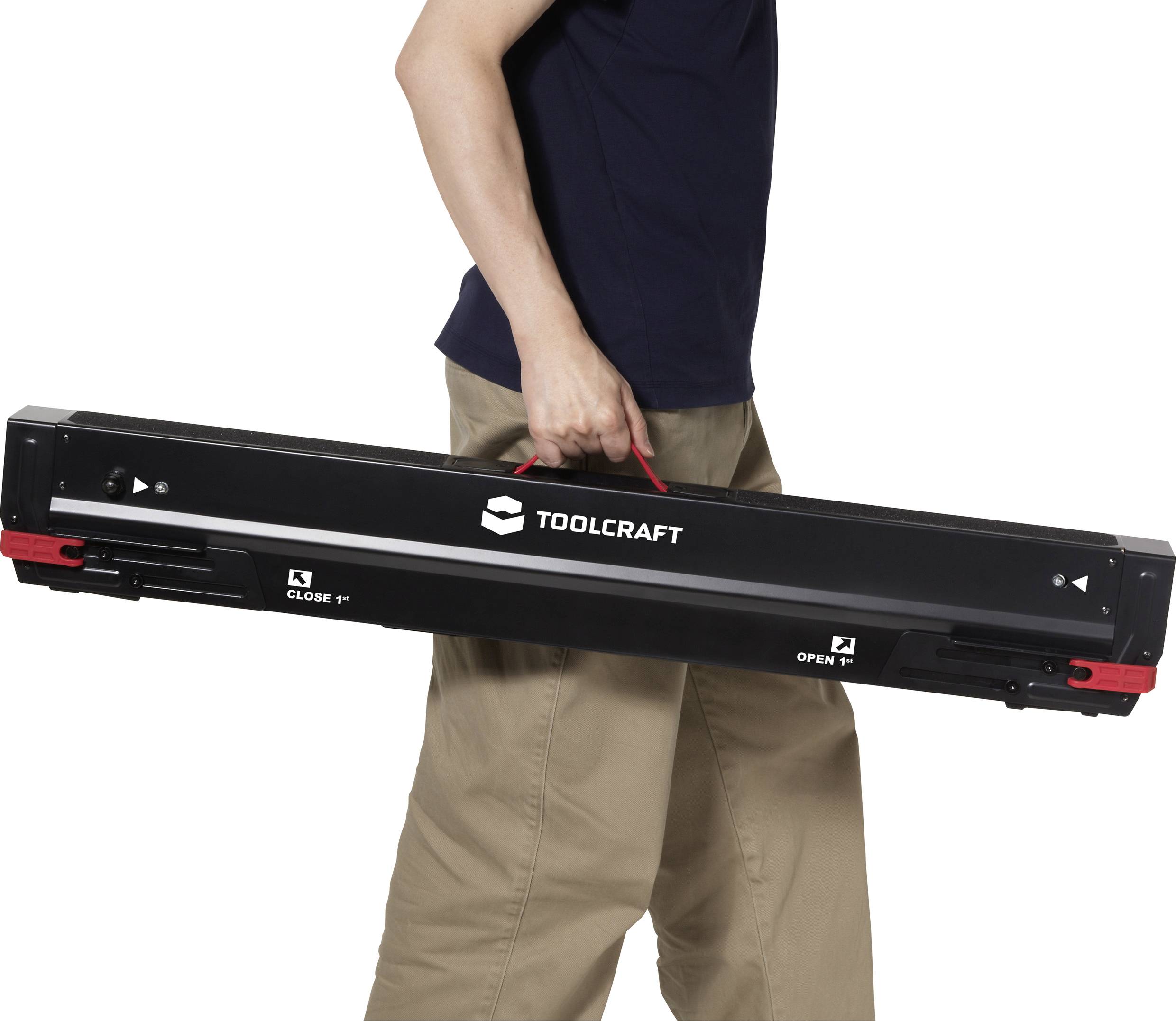 Eine Person in beigen Hosen und einem dunklen Hemd trägt eine lange schwarze Toolcraft-Werkzeugkiste mit einem Griff, die Anweisungen zum 