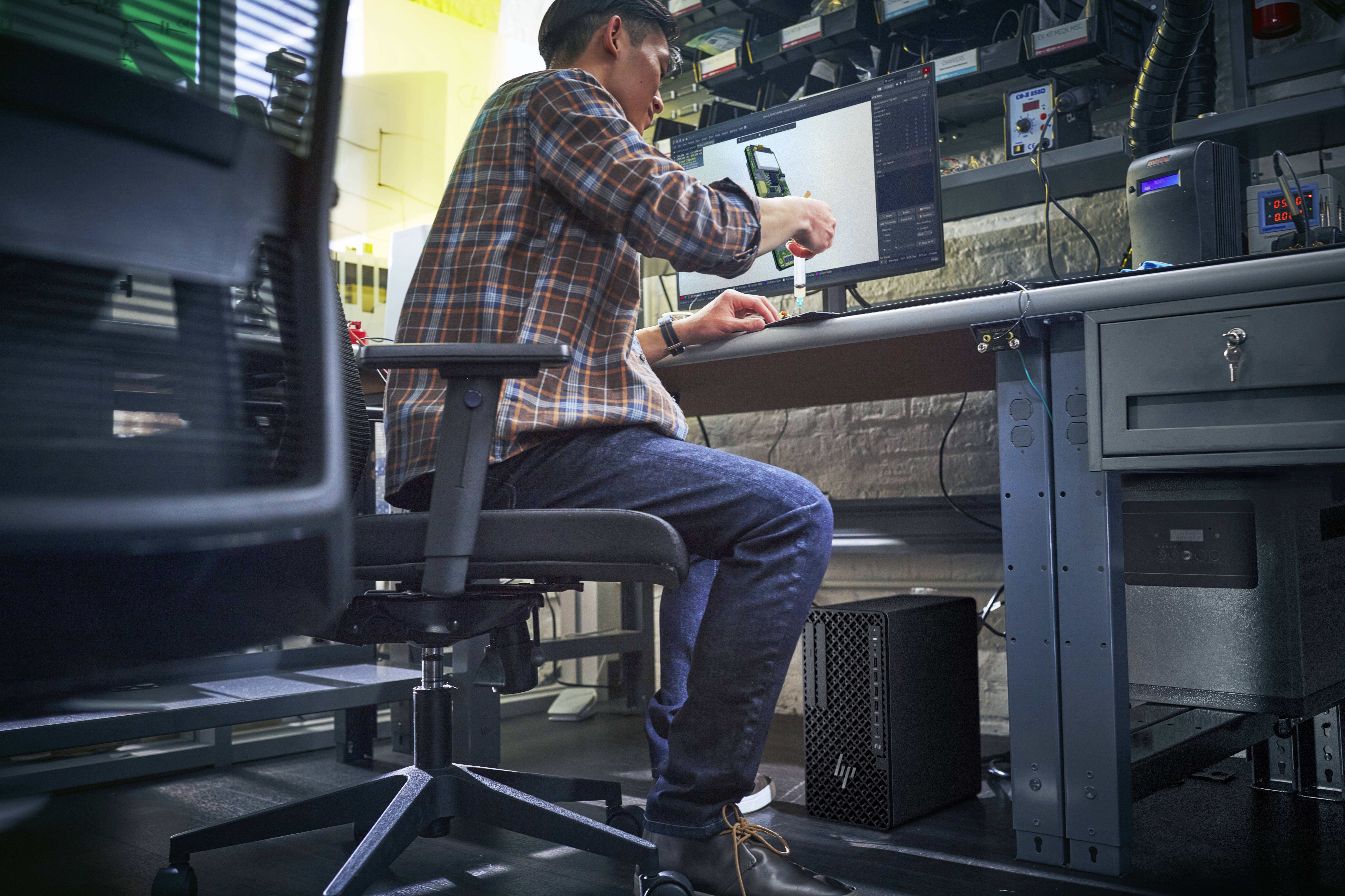 Eine Person, die an einem Schreibtisch sitzt und einen Computer benutzt, umgeben von elektronischer Ausrüstung und Werkzeugen in einer Werkstatt- oder Laborumgebung.