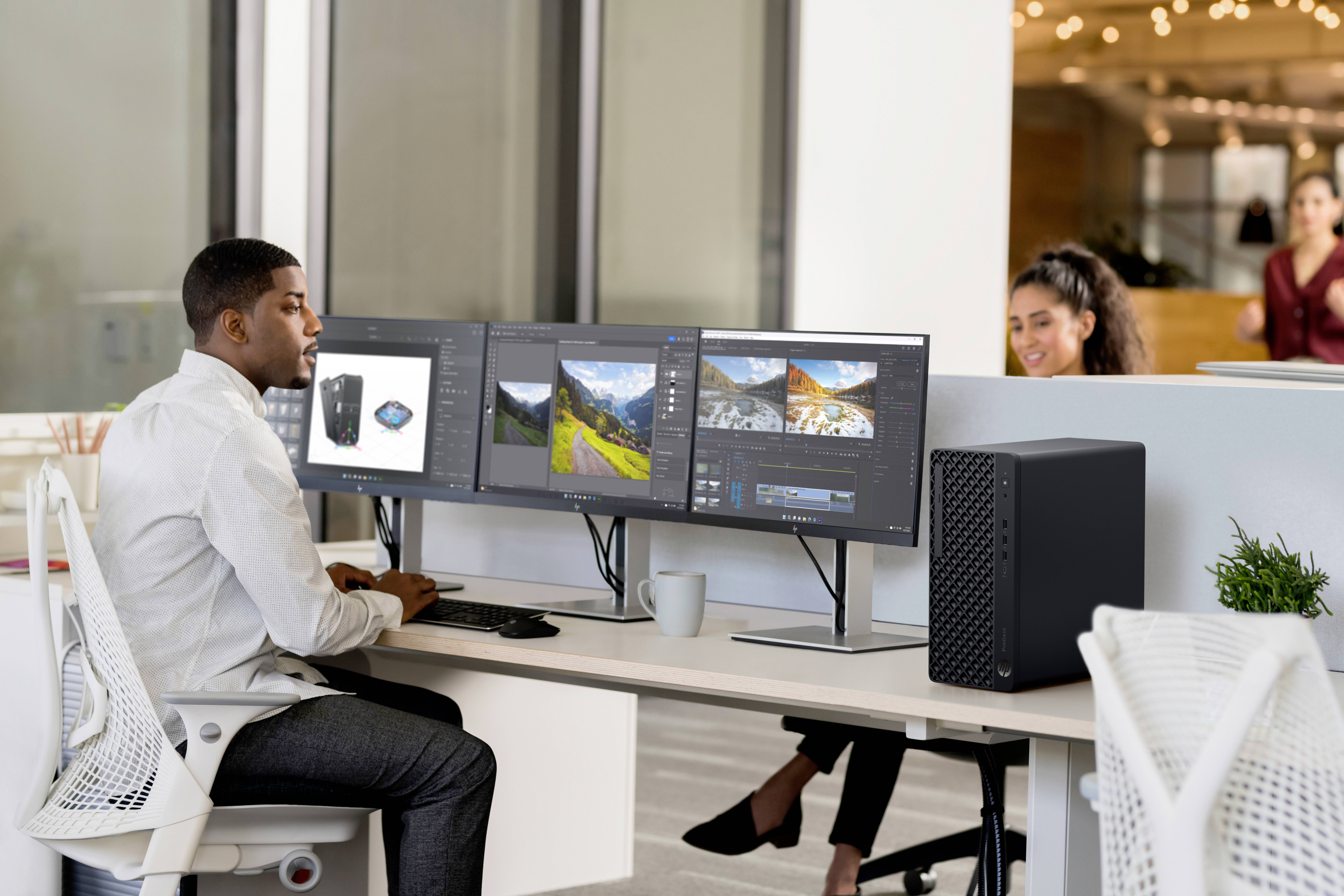 Eine Person arbeitet an einem Schreibtisch mit drei Computermonitoren, auf denen Designsoftware geöffnet ist. Im Hintergrund im Bürobereich ist eine weitere Person sichtbar.