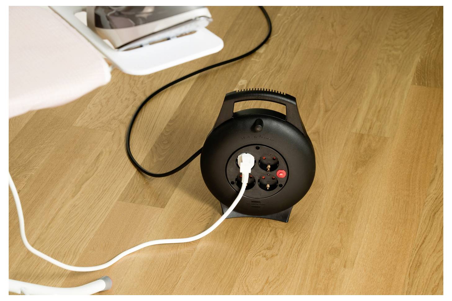 Schwarze Kabelrolle Verlängerungskabel mit mehreren Steckdosen auf einem Holzboden, ein weißer Stecker verbunden und eine rote Leuchte eingeschaltet.