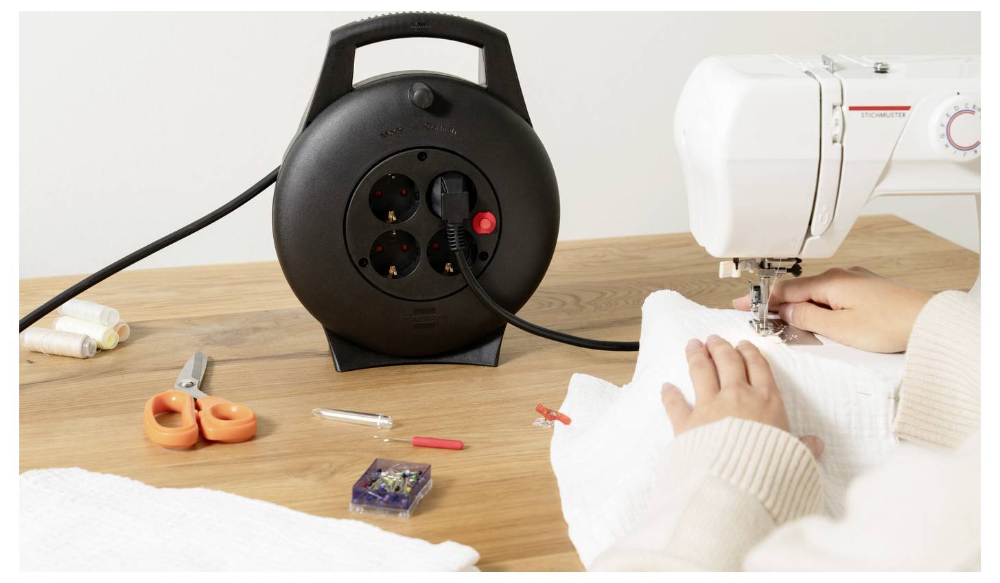 Eine Person näht Stoff an einer weißen Nähmaschine an einem Holztisch, mit Faden, Schere, Stecknadeln und einer schwarzen Verlängerungskabelrolle in der Nähe.