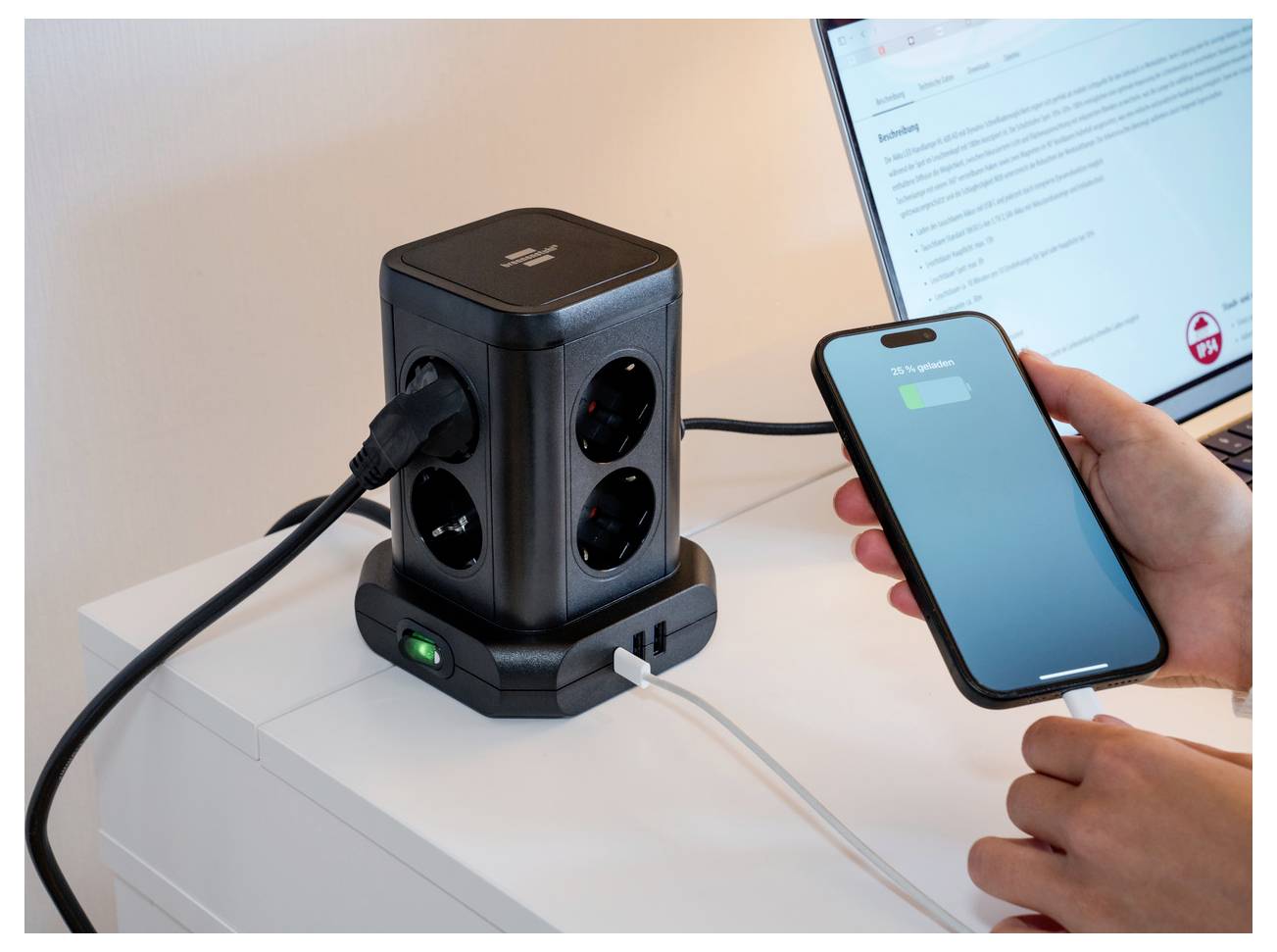 Hand, die ein Smartphone in eine würfelförmige Desktop-Stromstation mit mehreren AC-Steckdosen und Ladeanschlüssen neben einem Laptop einsteckt.