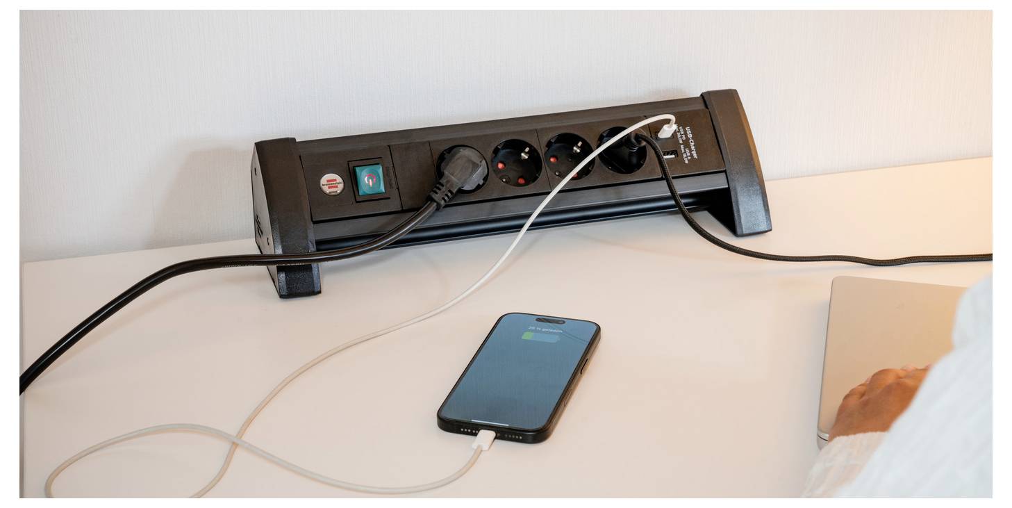 Ein Smartphone wird auf einem Schreibtisch über eine schwarze Steckerleiste mit mehreren Steckplätzen und Kabeln aufgeladen.