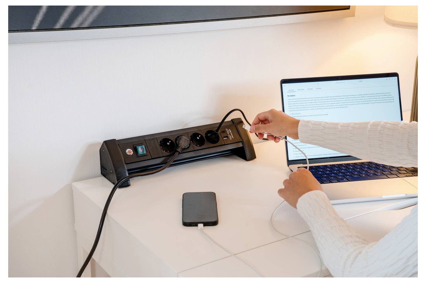 Eine Person steckt ein Kabel in eine Steckdosenleiste auf einem Schreibtisch neben einem Laptop und einer externen Festplatte auf einem weißen Tisch.