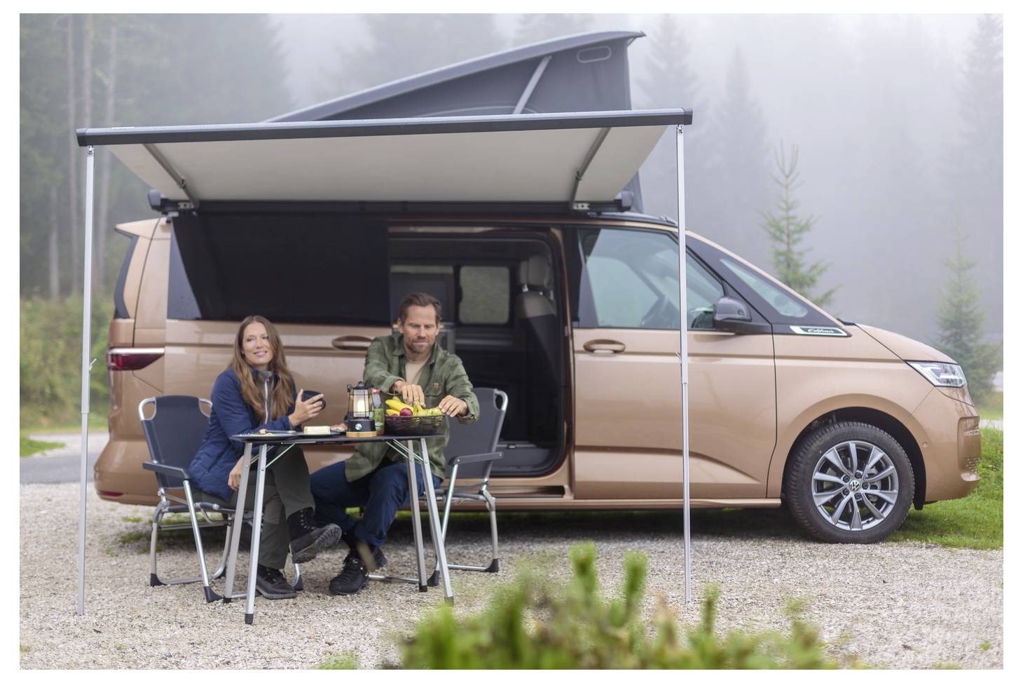 Ein Paar sitzt an einem Klapptisch unter einem aufklappbaren Sonnensegel neben einem beigen Wohnmobil auf einem nebligen Campingplatz im Freien.