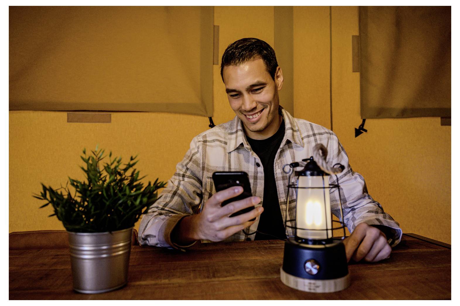 Ein lächelnder Mann an einem Tisch schaut auf sein Smartphone neben einer brennenden Laterne und einer Topfpflanze in einer warmen Innenumgebung.