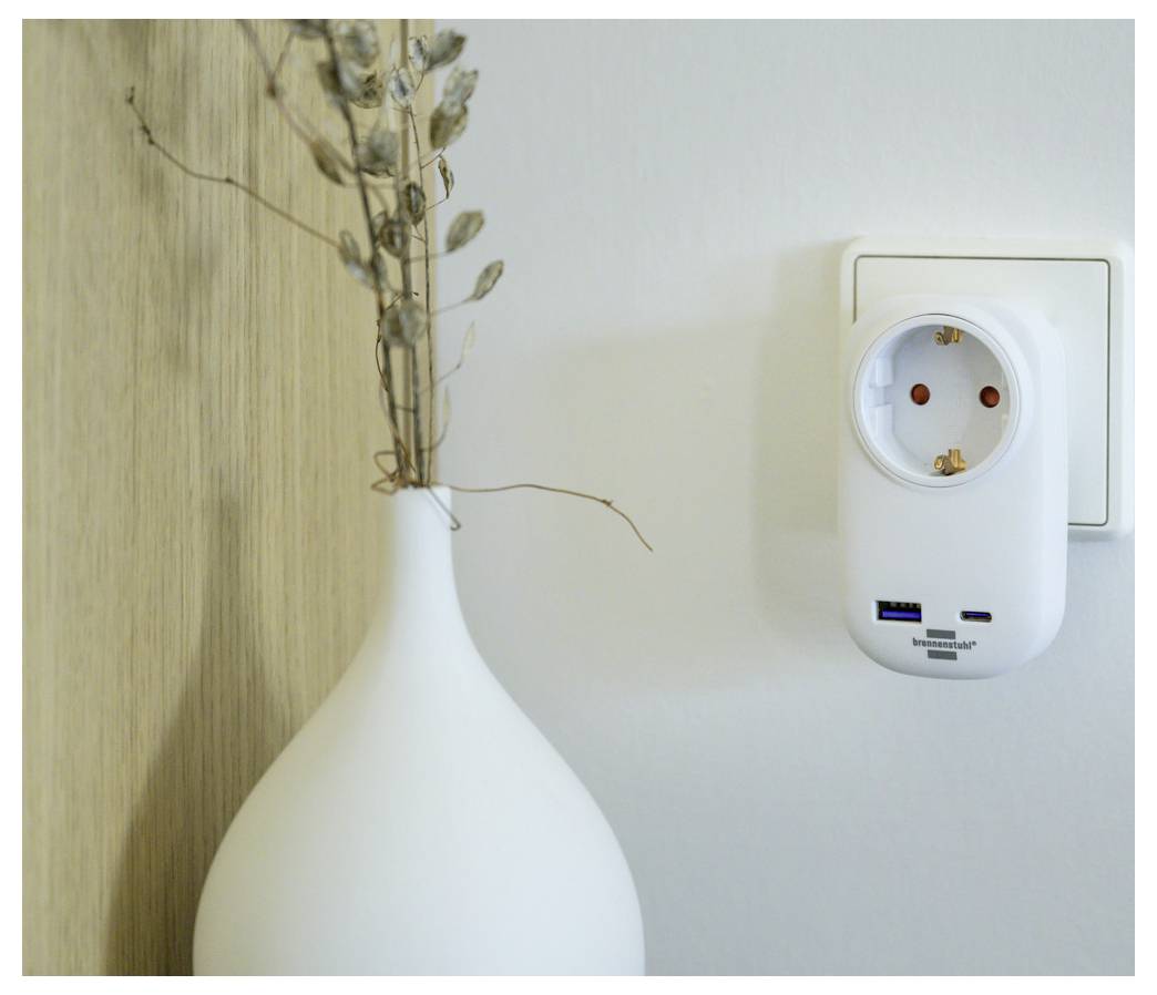 Weiße Vase mit getrockneten Stielen neben einer Wandsteckdose und einem Steckergerät an einer einfachen Wand.